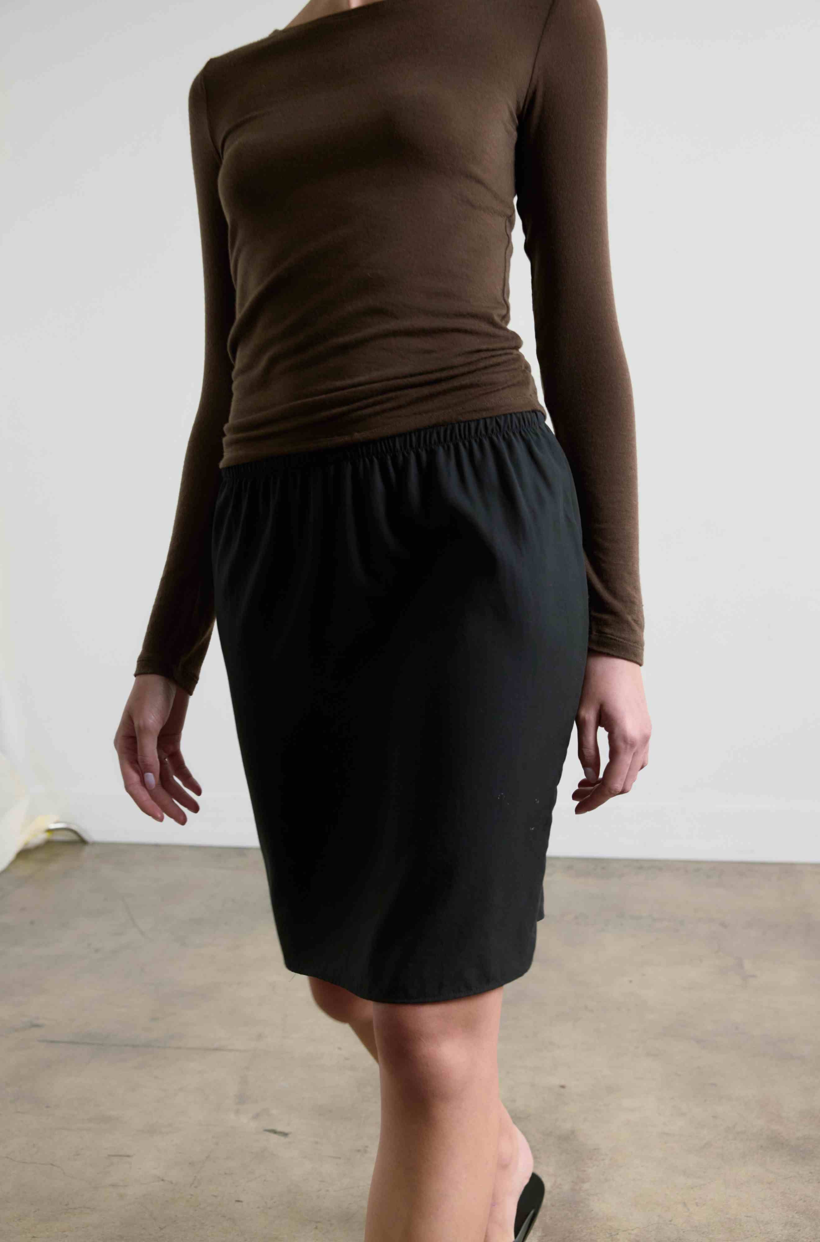 Person wearing a brown long-sleeve top and black skirt against a white wall.