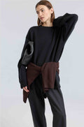 Person wearing a black sweater with a brown sweater wrapped around her waist and black pants on a light gray background