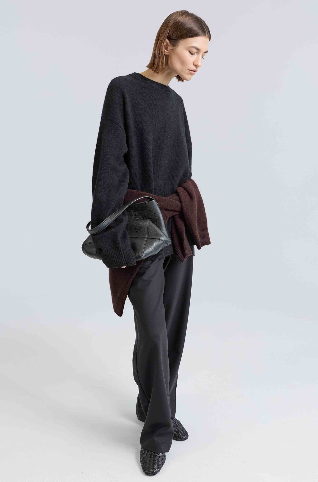 Person wearing a black sweater and gray pants holding a brown jacket on a light gray background