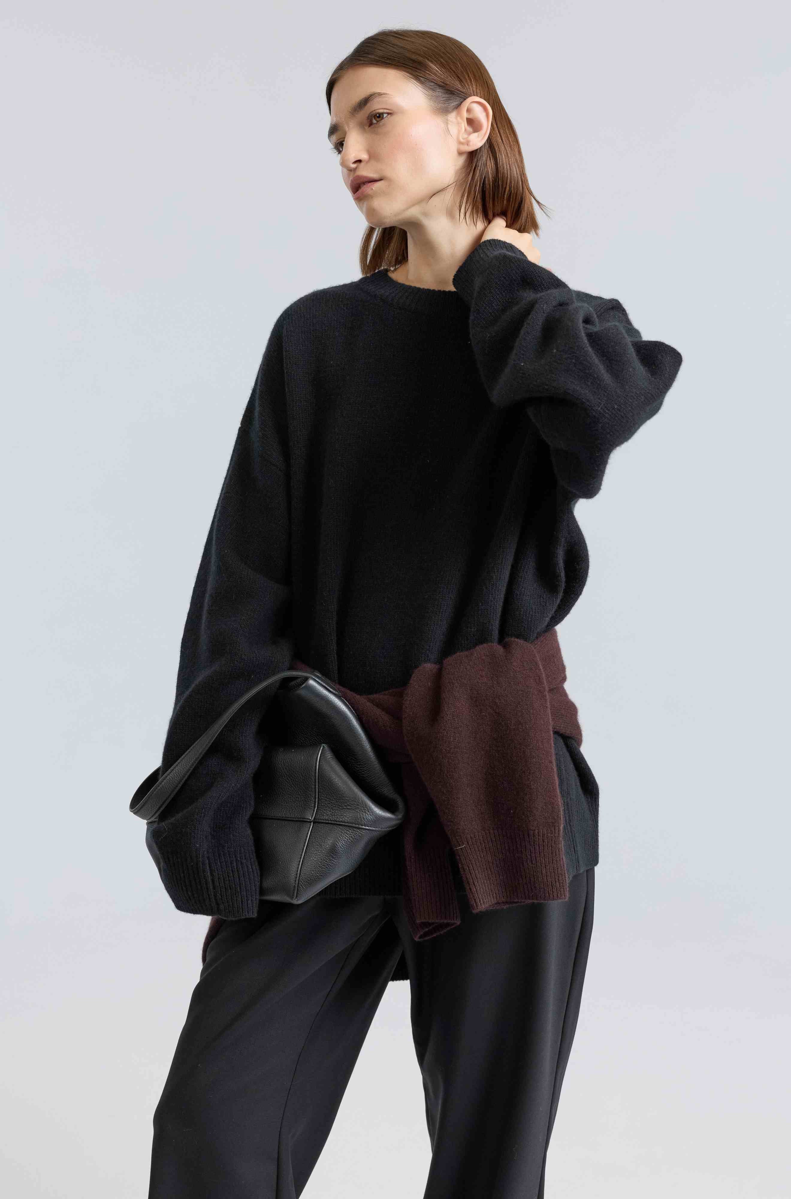 Woman wearing a black oversized sweater with a brown sweater tied around her waist on a light gray background