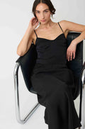 Woman in a black silk top and matching black silk skirt sitting on a modern chair against a white background