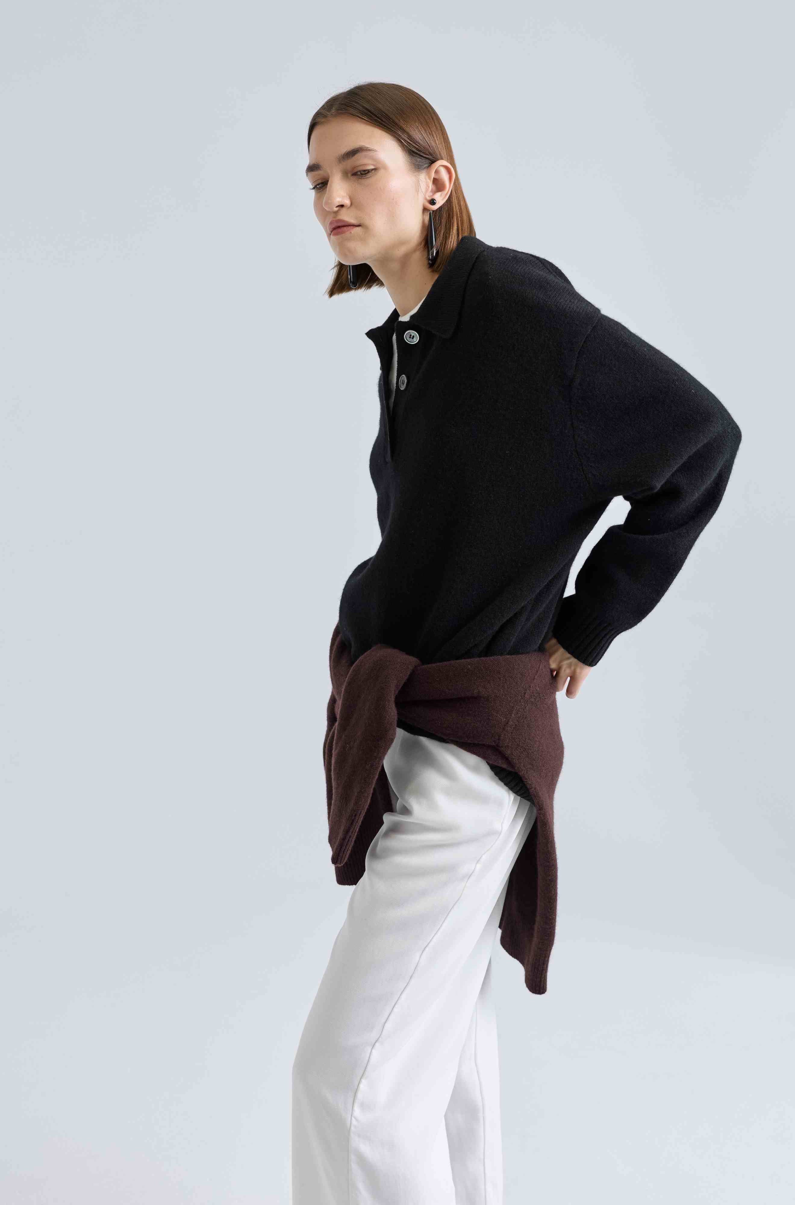 Person wearing a black sweater with a brown shawl over white pants on a light gray background