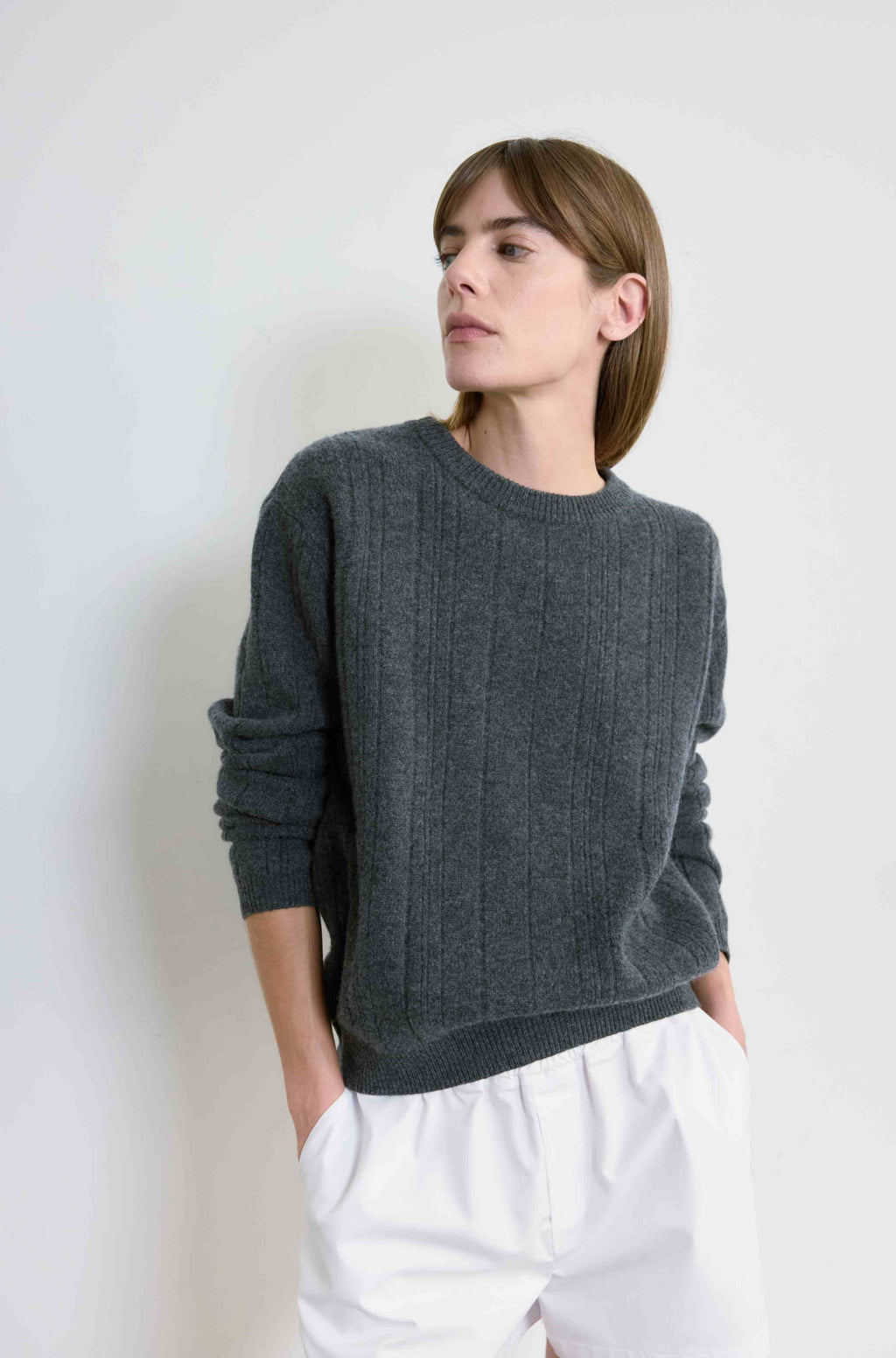 Woman wearing a dark gray sweater and white pants against a light background