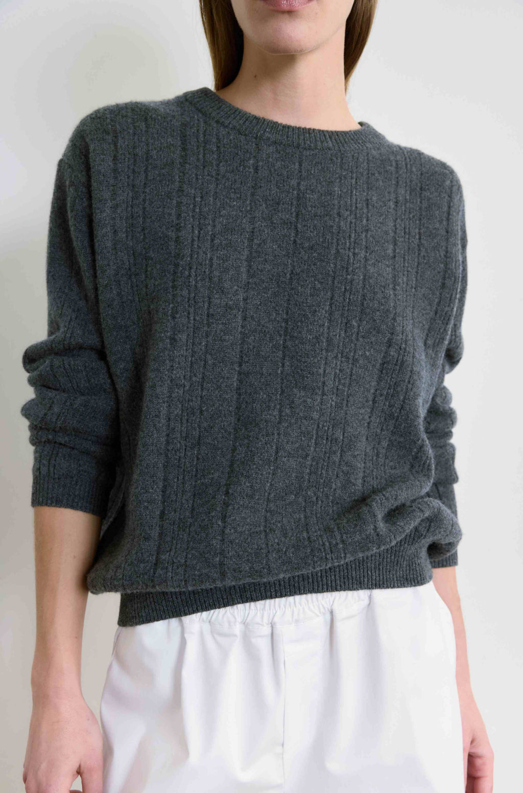 Person wearing a dark gray cable knit sweater and white pants on a plain background