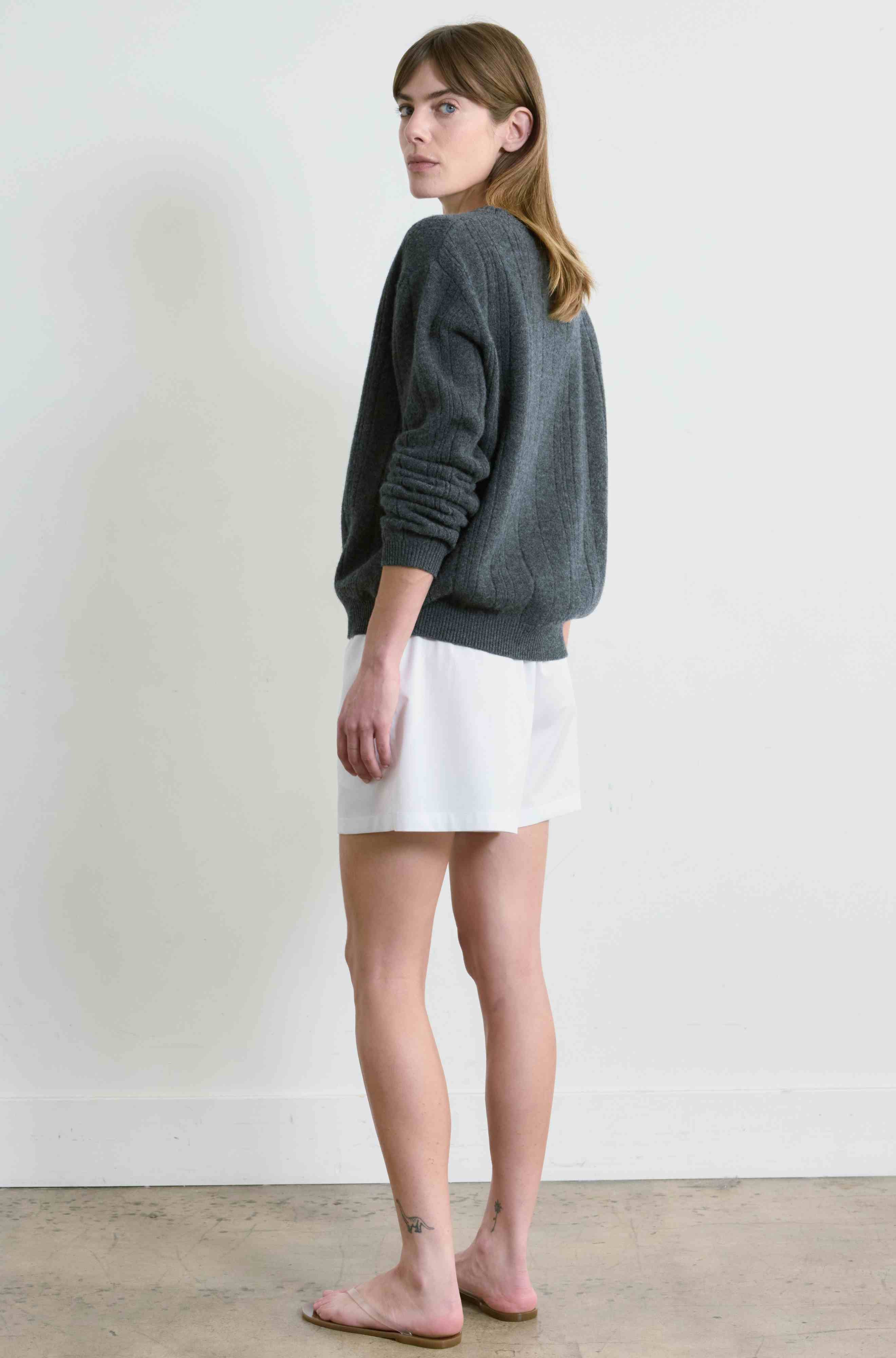 Woman wearing a gray sweater and white shorts standing against a plain white wall.