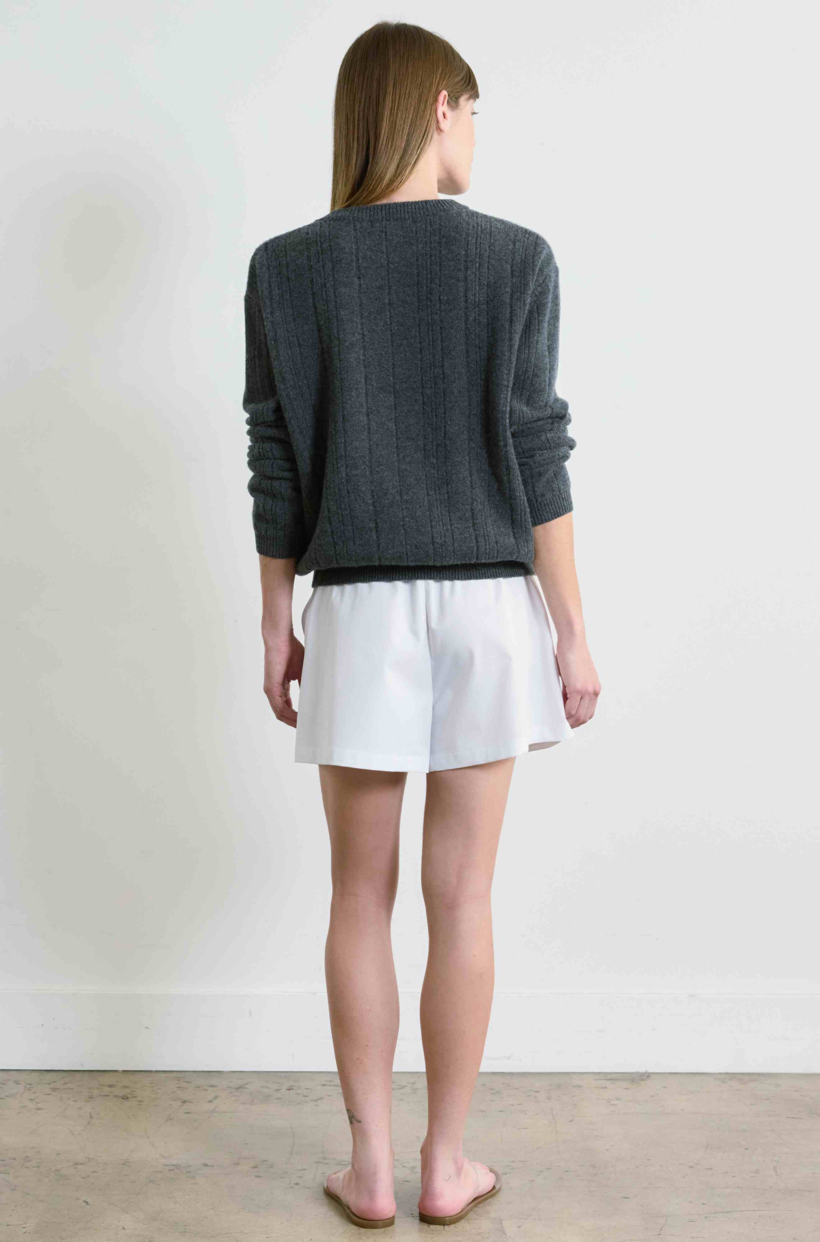 Person wearing a gray sweater and white shorts standing against a plain white wall.
