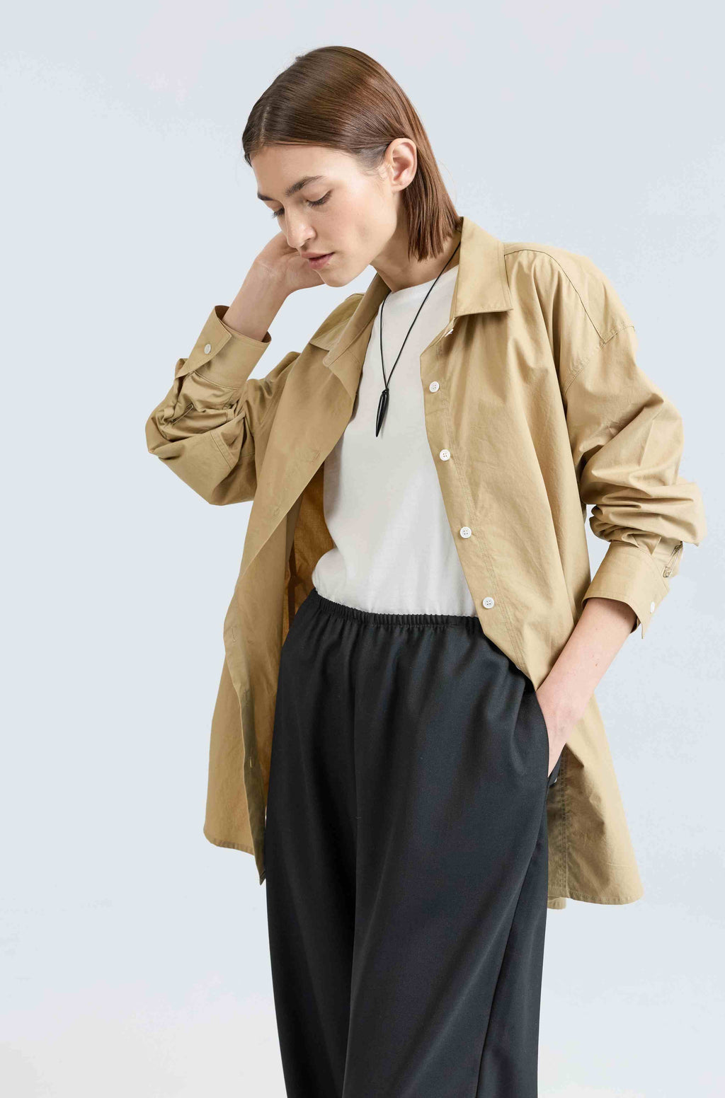 Woman wearing a beige button up shirt, white t-shirt, and black pants on a light gray background
