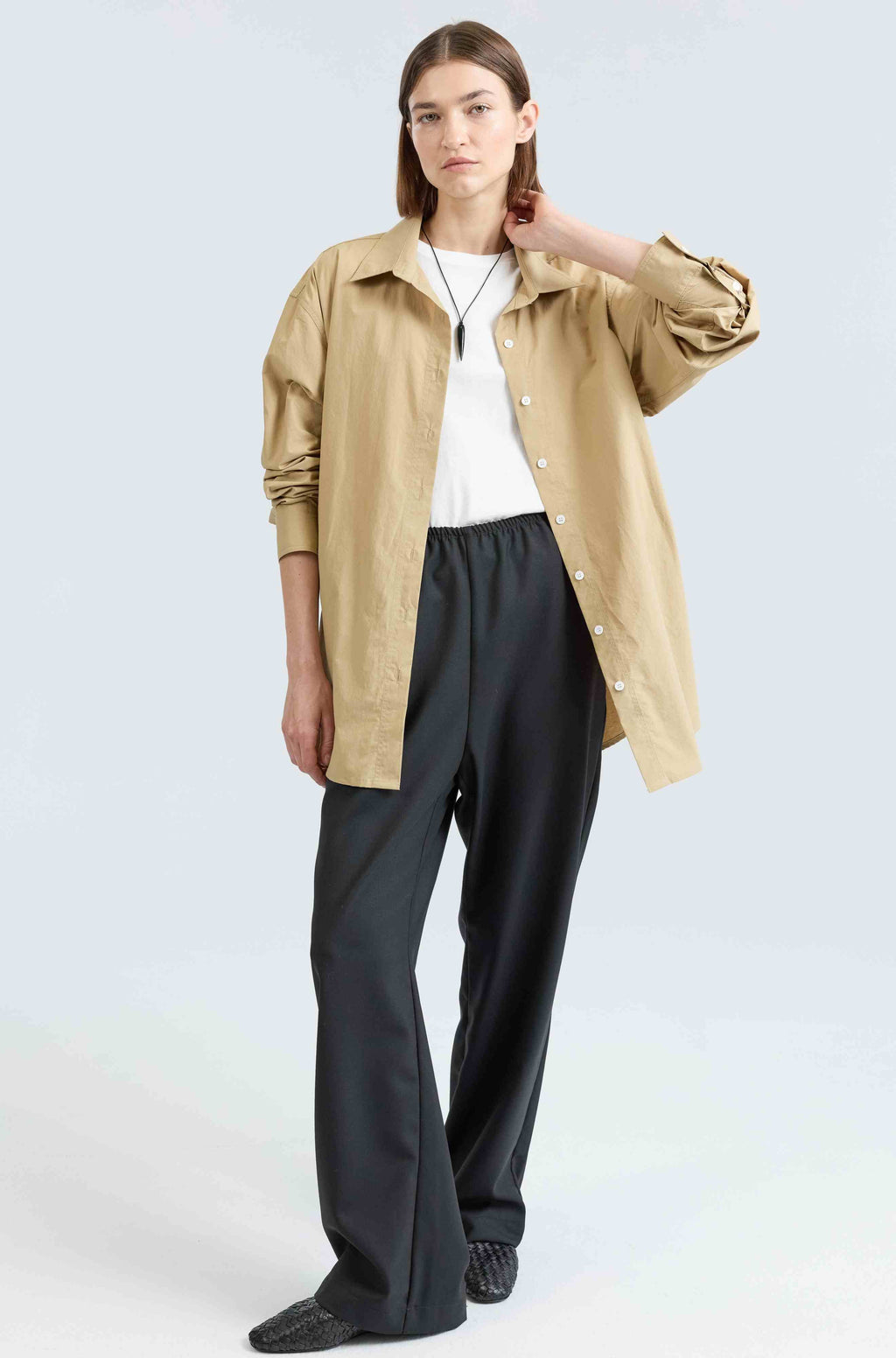 Woman wearing a beige button up shirt, white t-shirt, and black pants on a light gray background