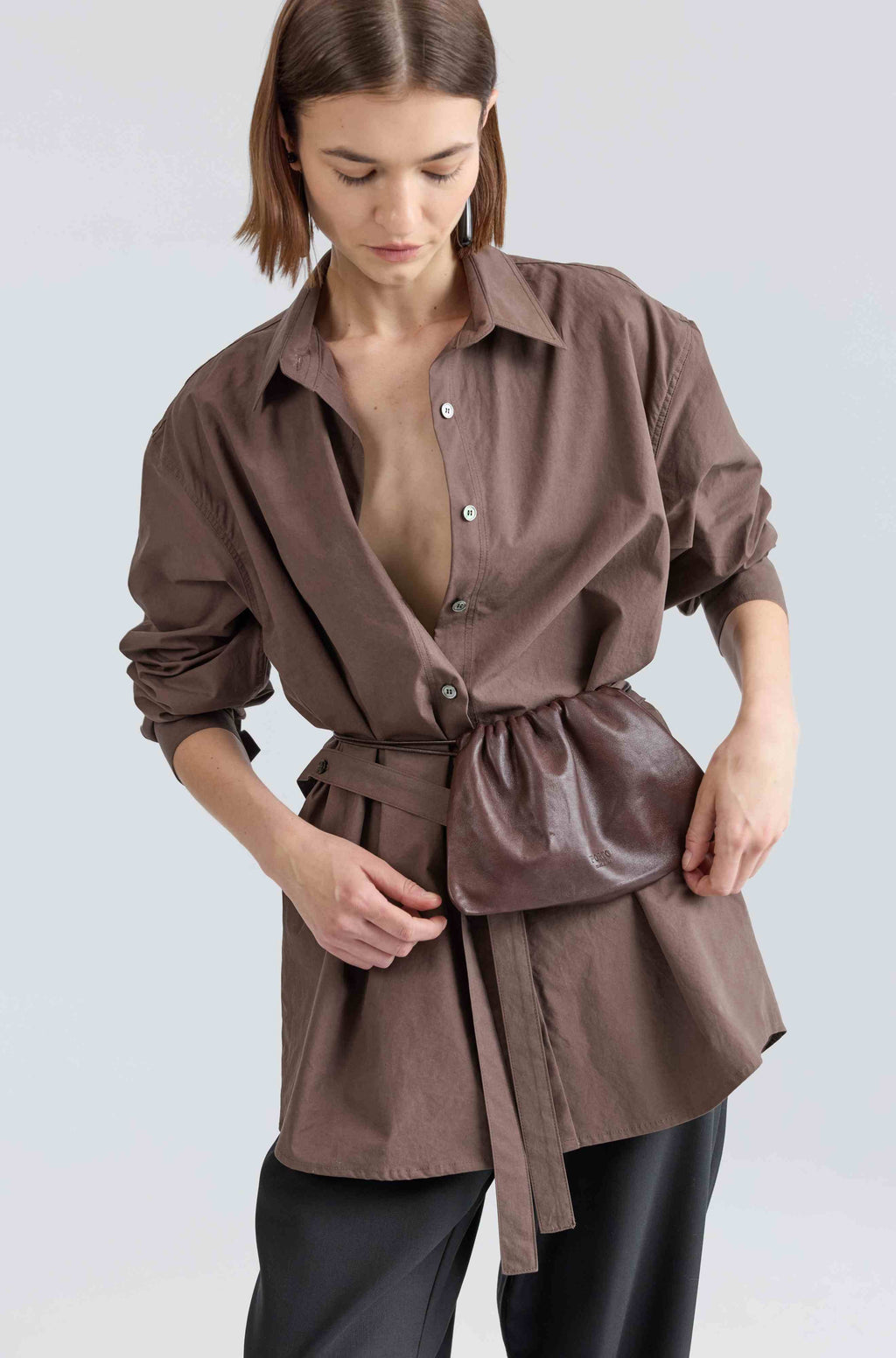 Woman wearing a brown belted shirt over black pants on a light gray background