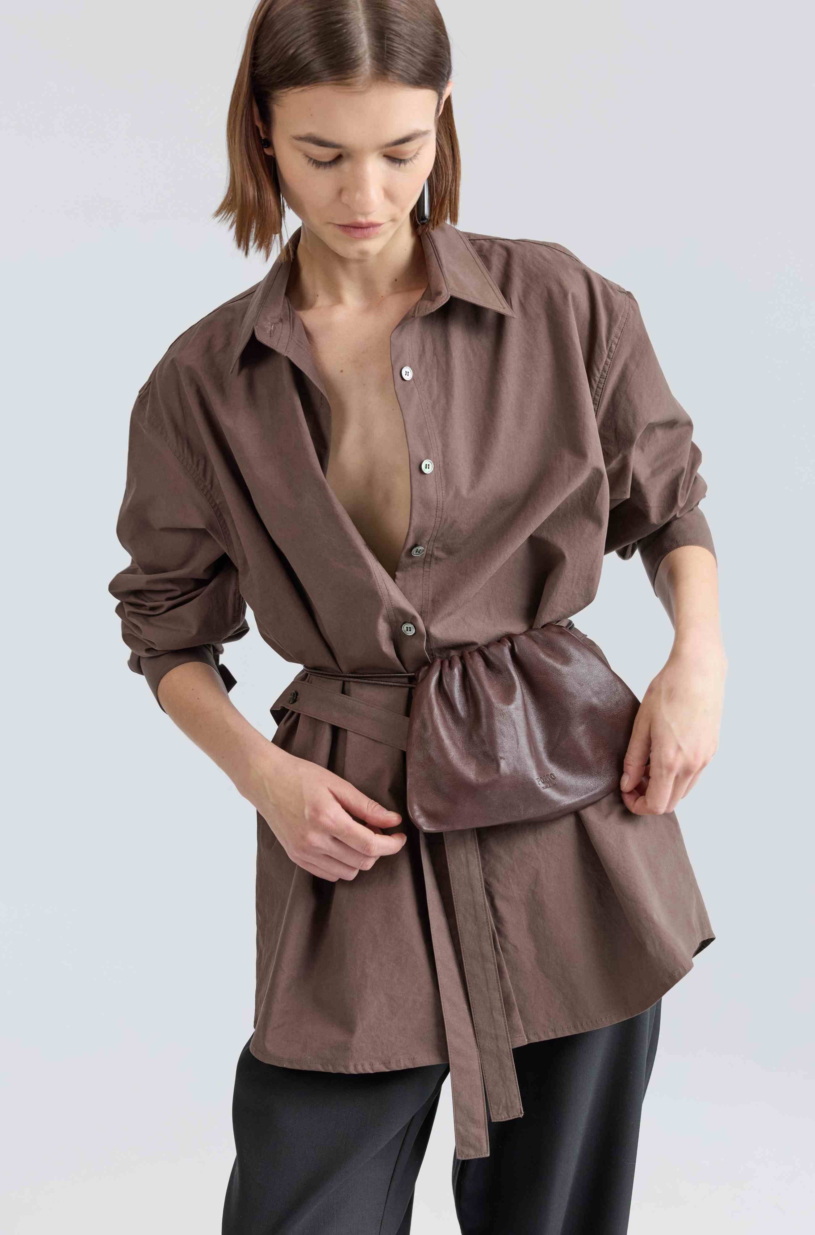 Woman wearing a brown belted shirt over black pants on a light gray background