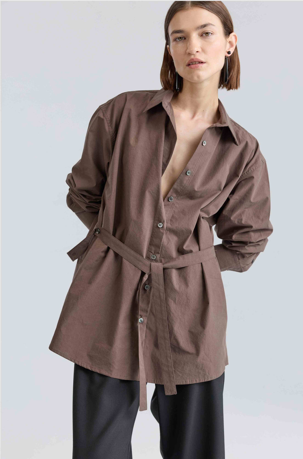Woman wearing a brown belted shirt over black pants on a light gray background