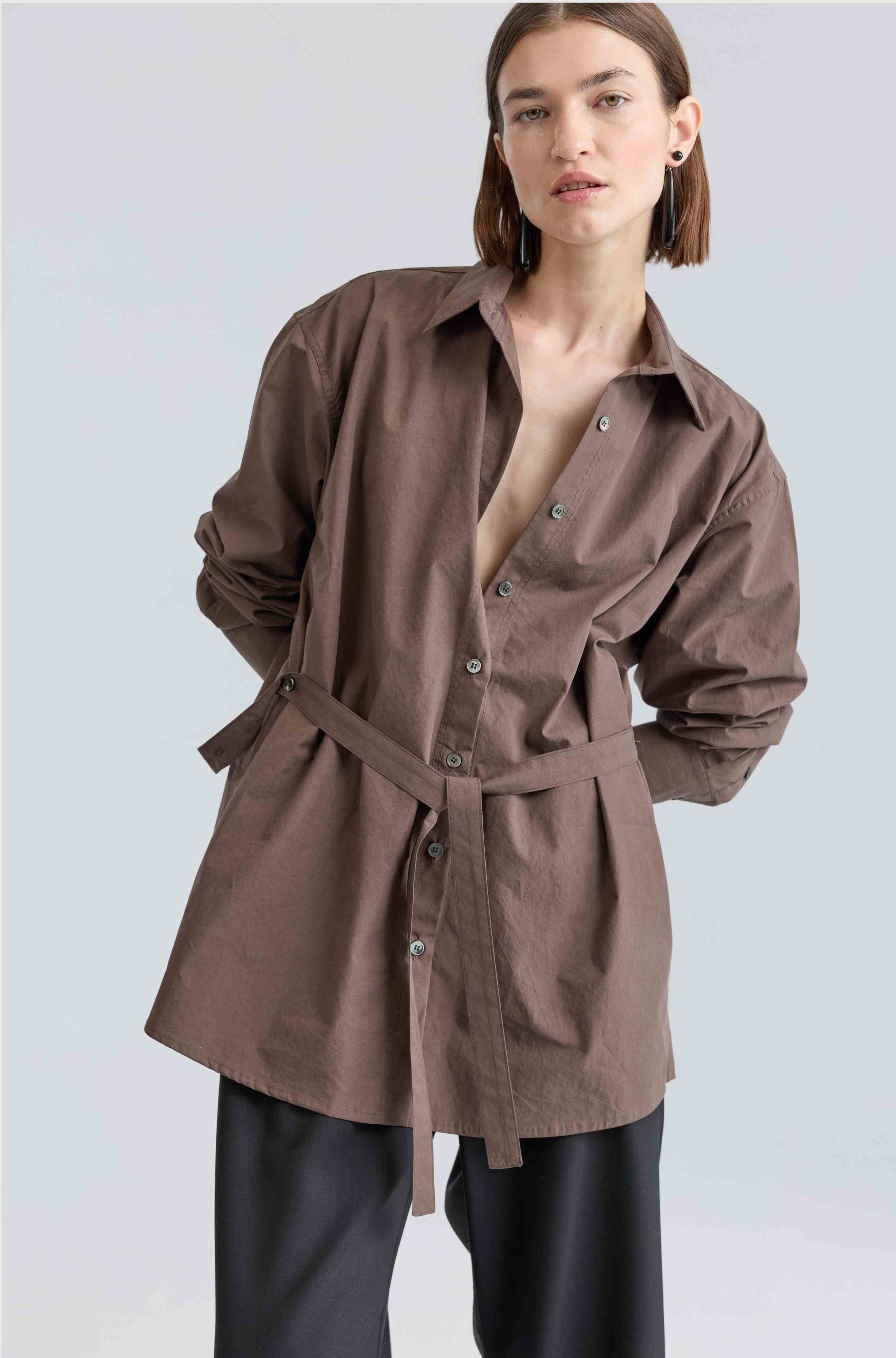 Woman wearing a brown belted shirt over black pants on a light gray background
