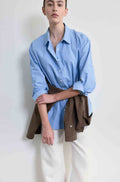 Person wearing a light blue shirt and white pants with a brown jacket over the waist on a plain background