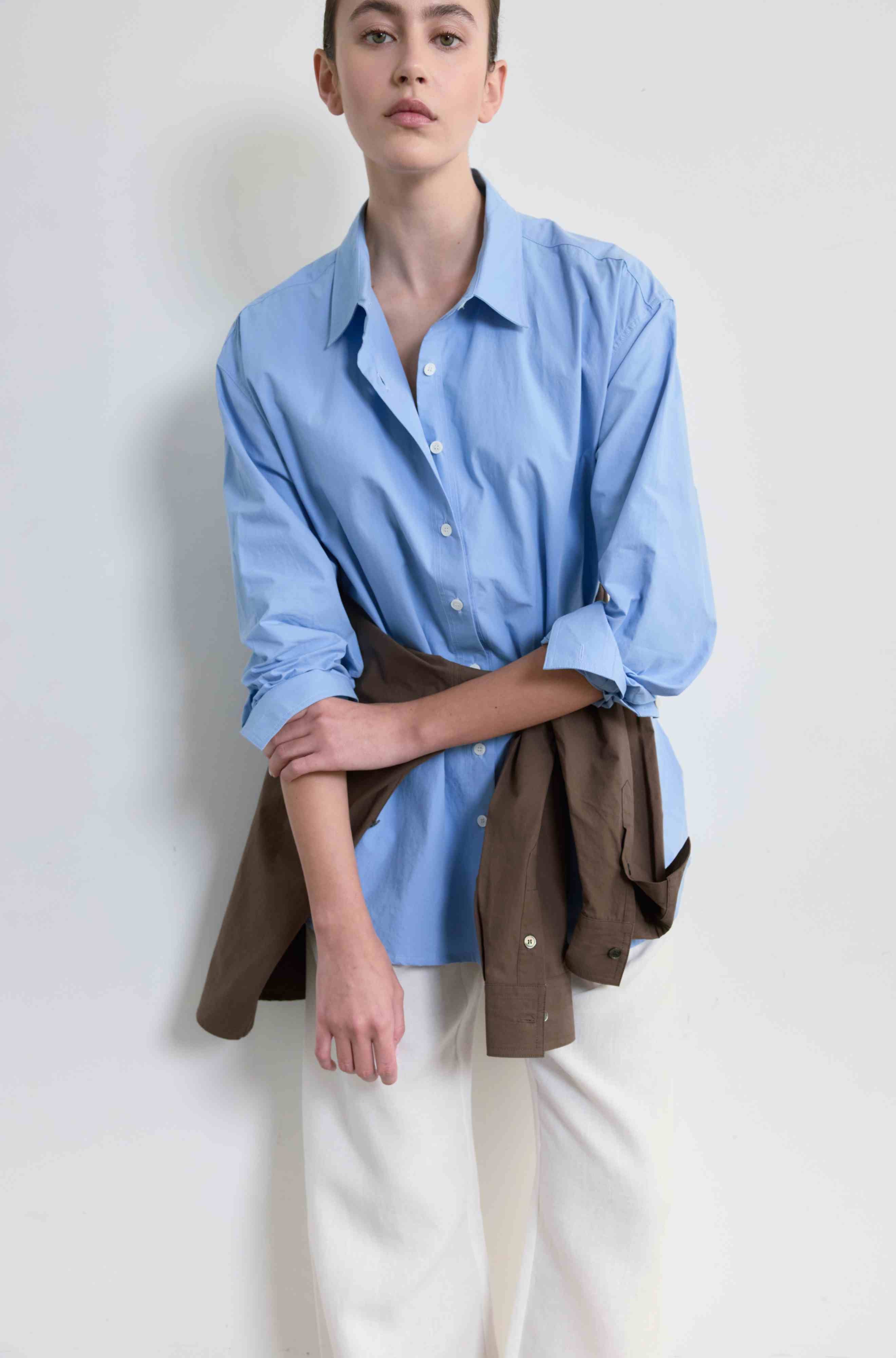 Person wearing a light blue shirt and white pants with a brown jacket over the waist on a plain background
