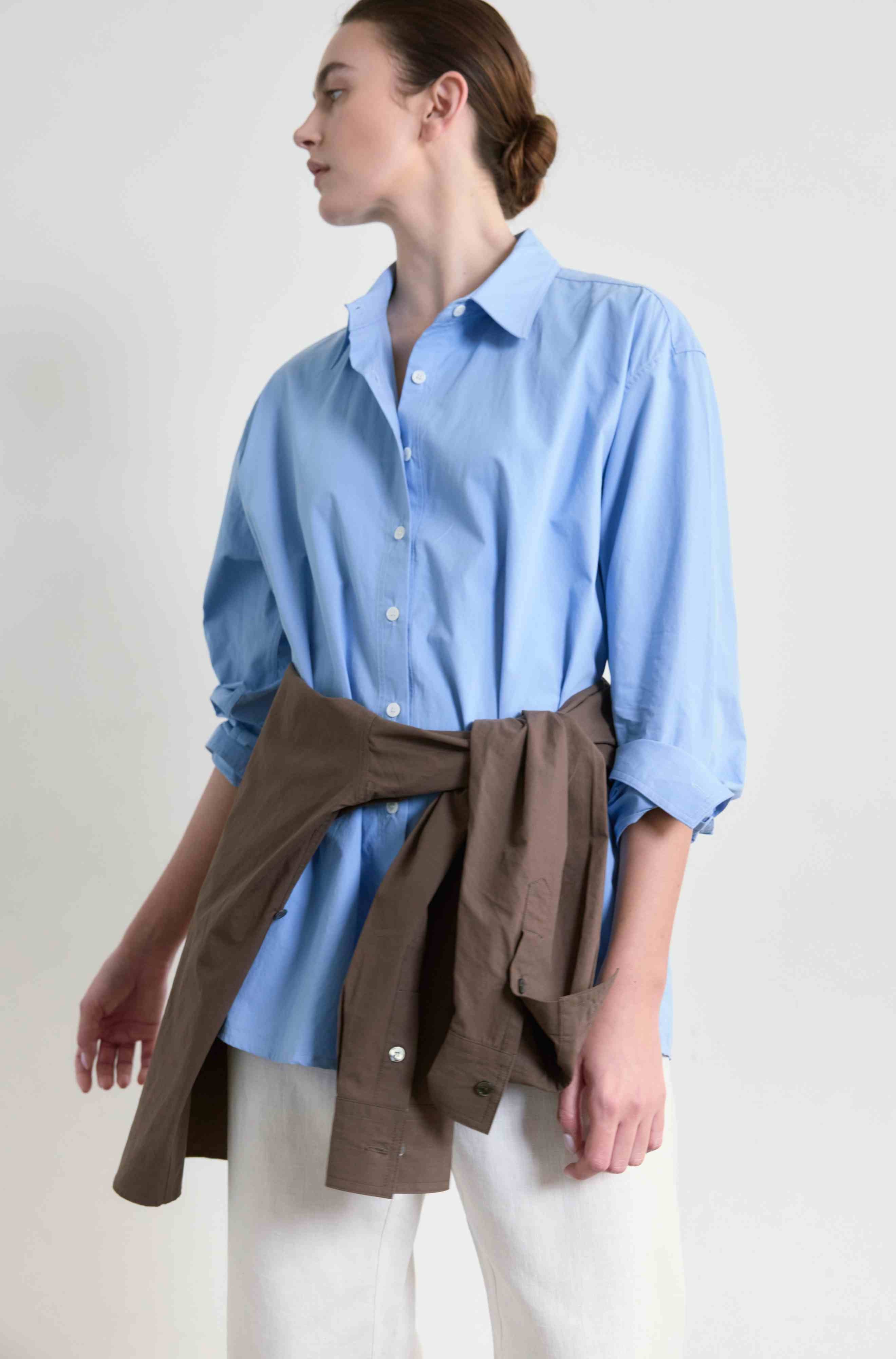 Woman wearing a light blue shirt with rolled-up sleeves and a brown jacket around her waist on a white background