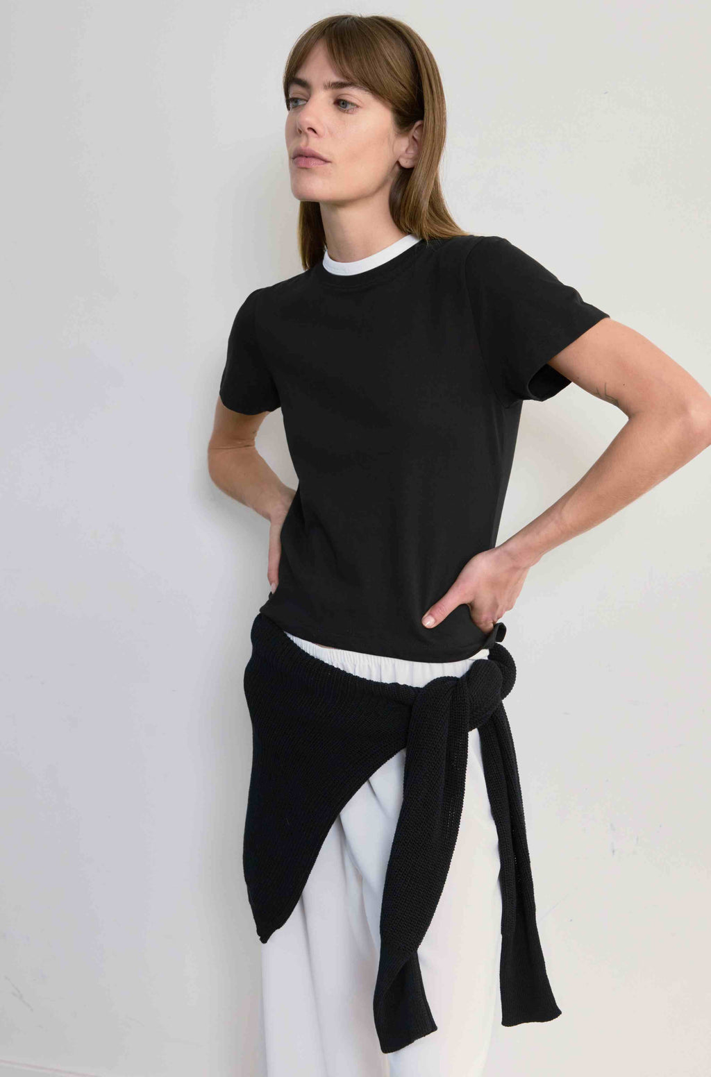 Woman wearing a black top and white pants with a black scarf tied around her waist against a plain background