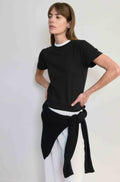 Woman wearing a black top and white pants with a black scarf tied around her waist against a plain background