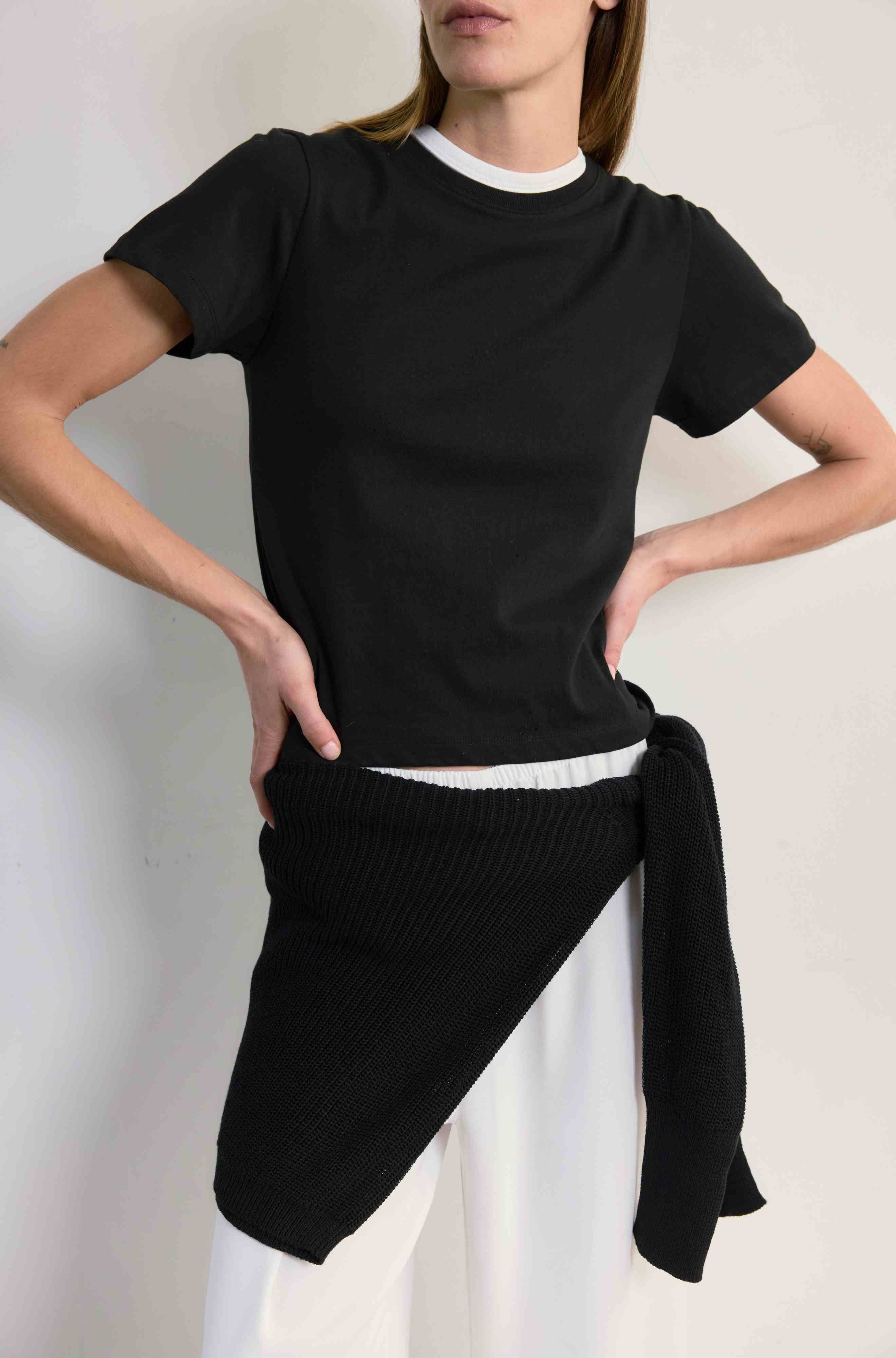 Person wearing a black t-shirt and white pants with a black sweater tied around the waist against a plain background
