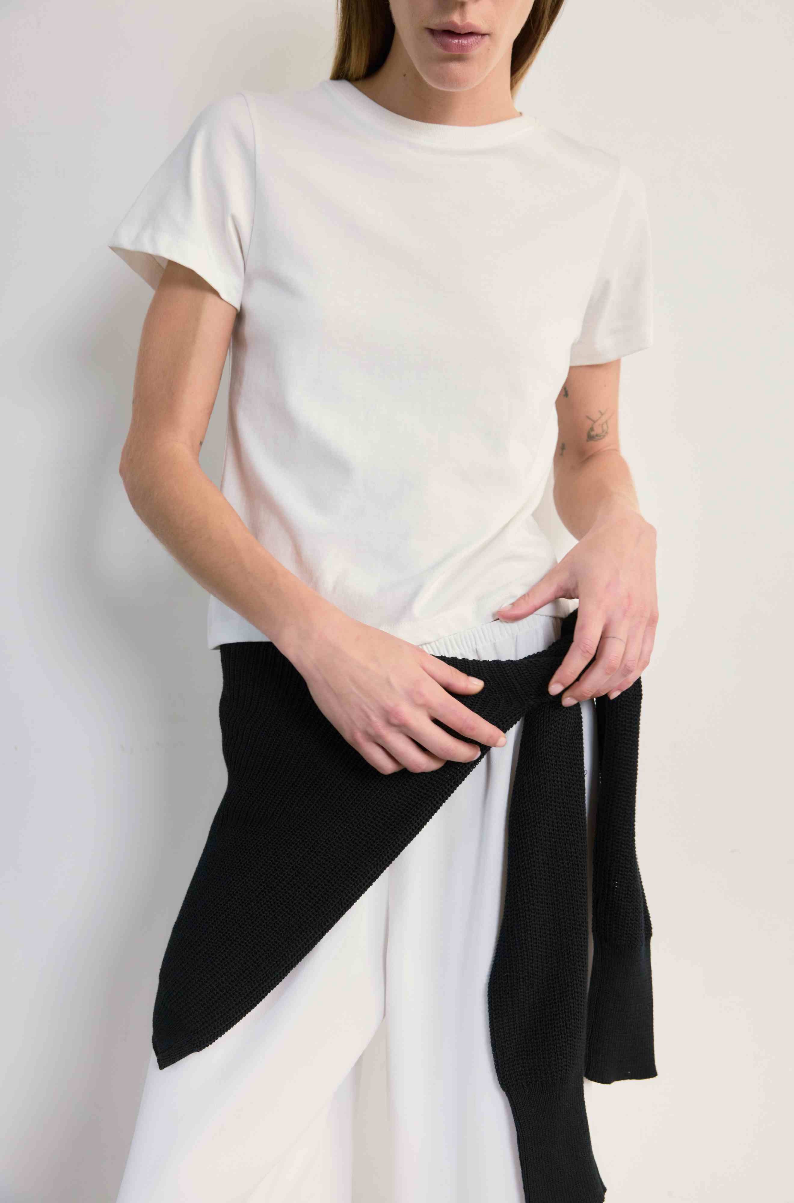 Person wearing a white t-shirt and black sweater tied around her waist against a plain background