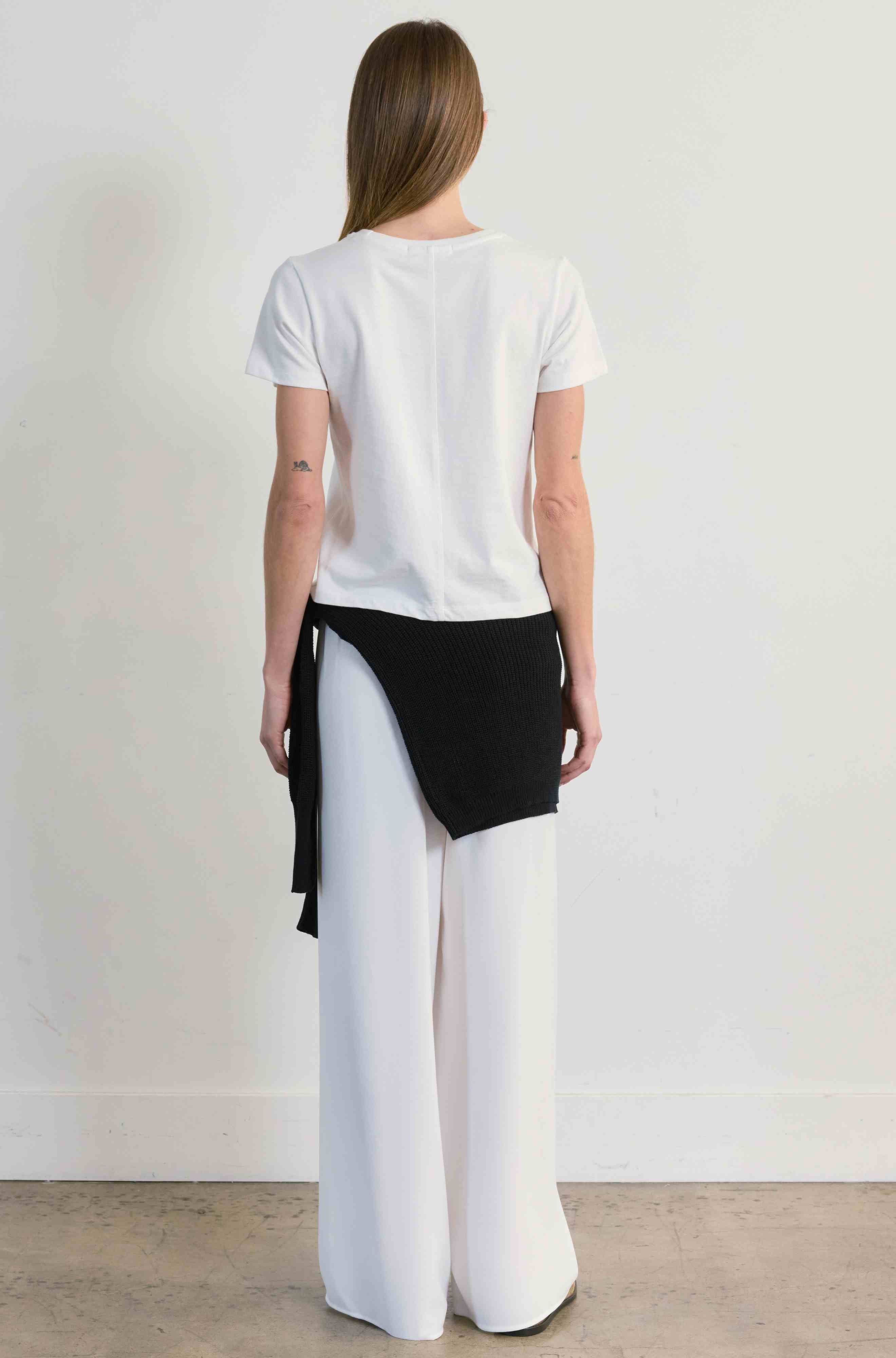 Person wearing a white t-shirt and white pants with a black sweater around her waist, standing against a plain background.