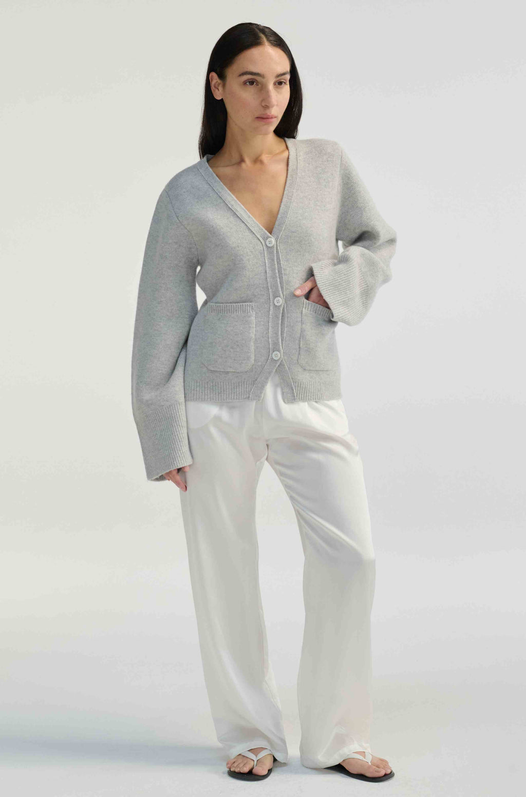 Woman wearing a light gray cardigan and white silk pants on a plain background