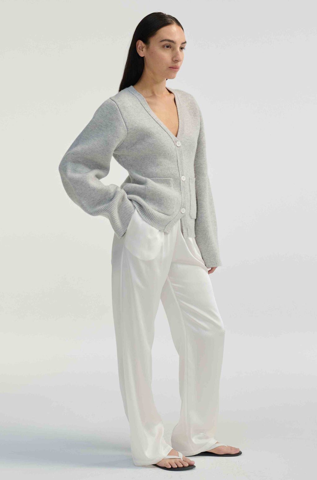 Woman wearing a gray cardigan and white silk pants on a plain background
