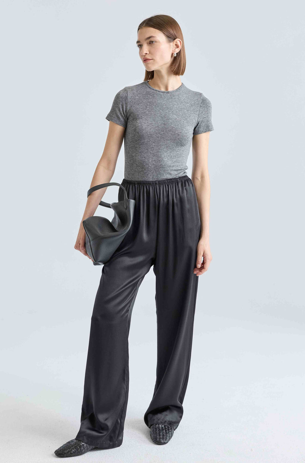 Woman wearing a gray t-shirt and black pants holding a black handbag on a light gray background
