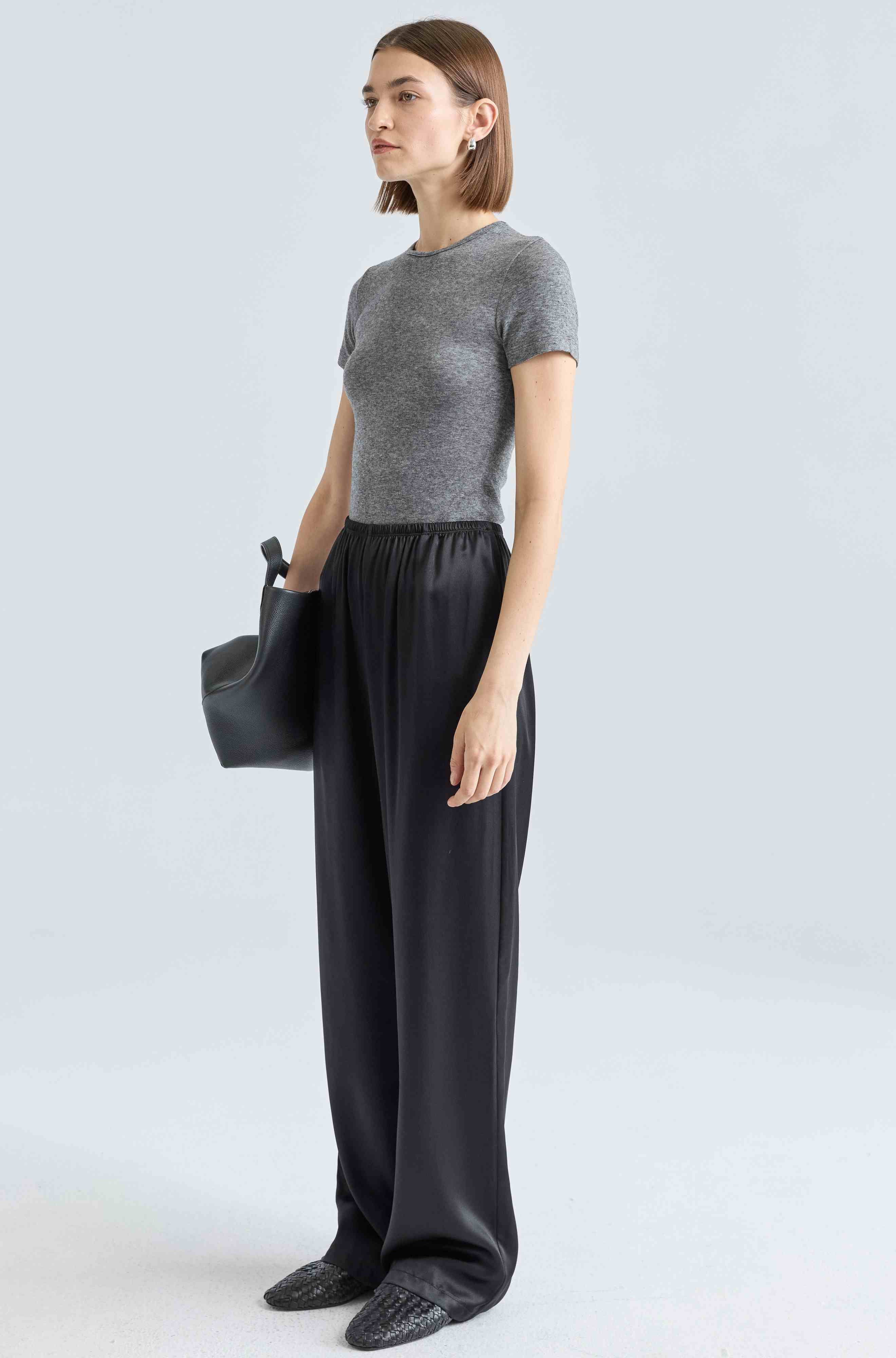 Woman wearing a gray sweater and black pants holding a black bag on a white background
