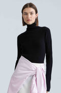 Woman wearing a black turtleneck sweater and a light pink shirt tied around her waist against a light gray background