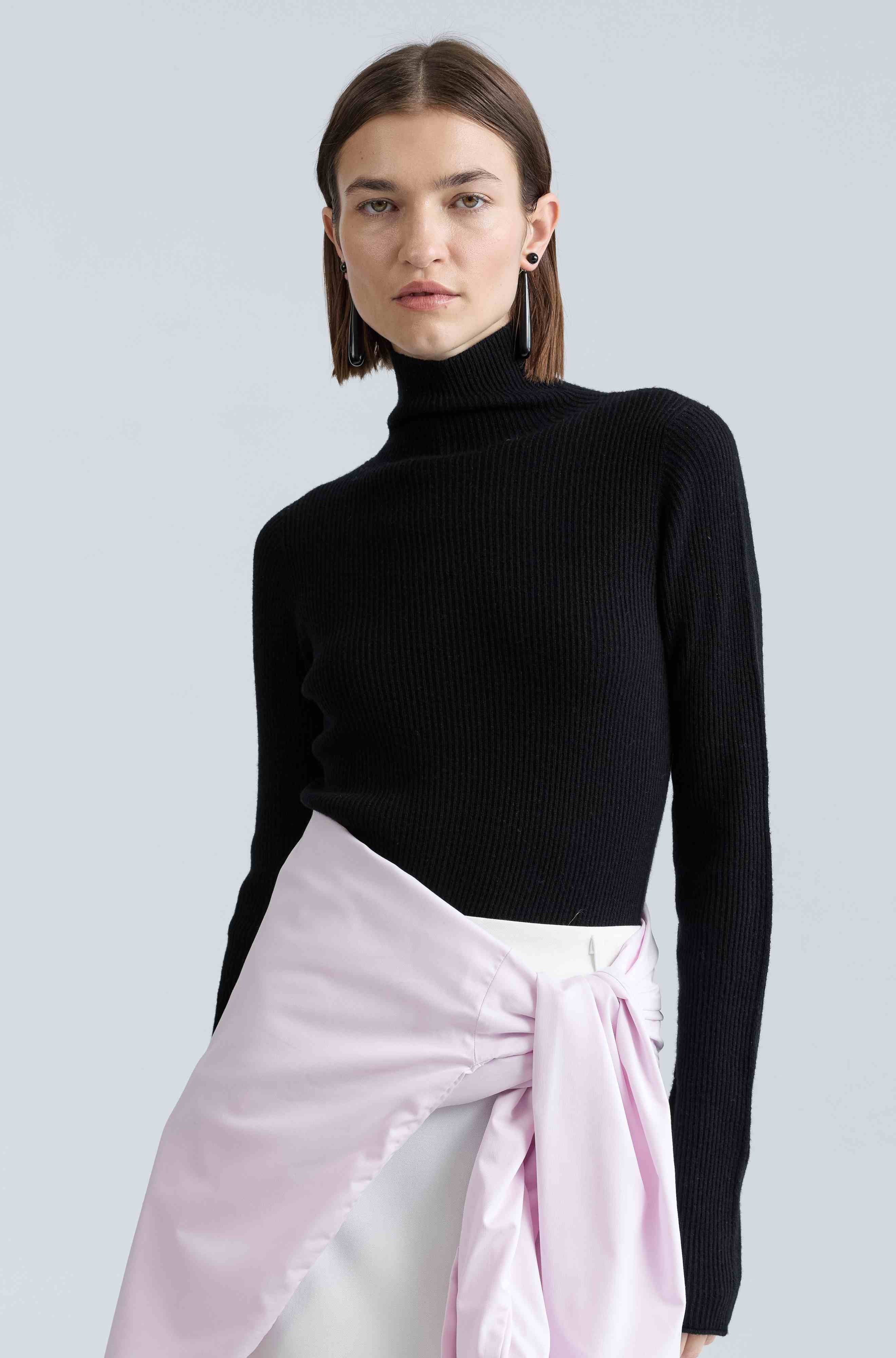 Woman wearing a black turtleneck sweater and a light pink shirt tied around her waist against a light gray background