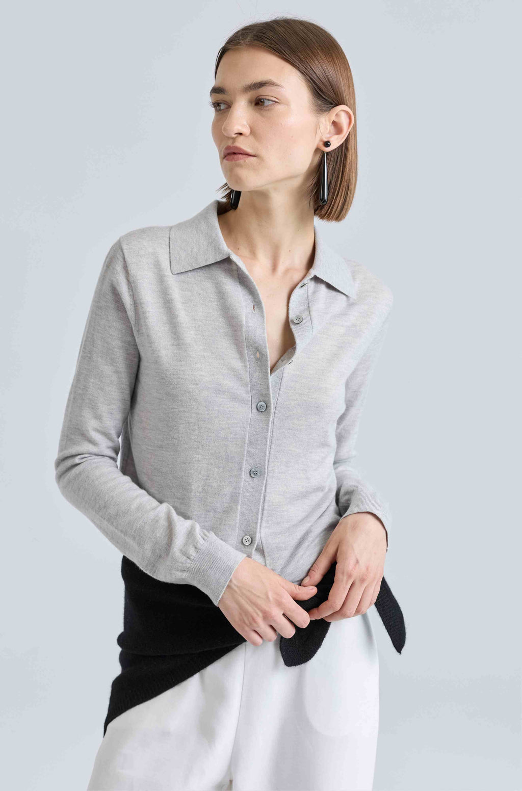 Woman wearing a gray button-up shirt over white pants and a black scarf tied around her waist on a light gray background