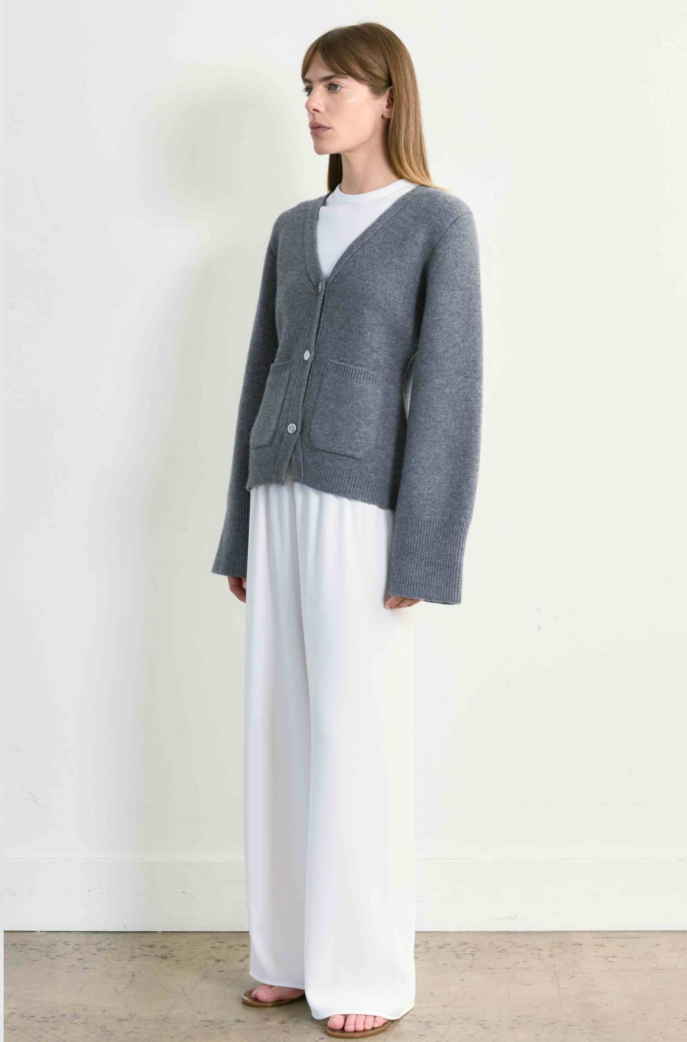 Woman wearing a gray cardigan over a white top and pants on a plain background