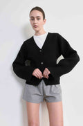 Person wearing a black cardigan over a white shirt and plaid shorts on a light background