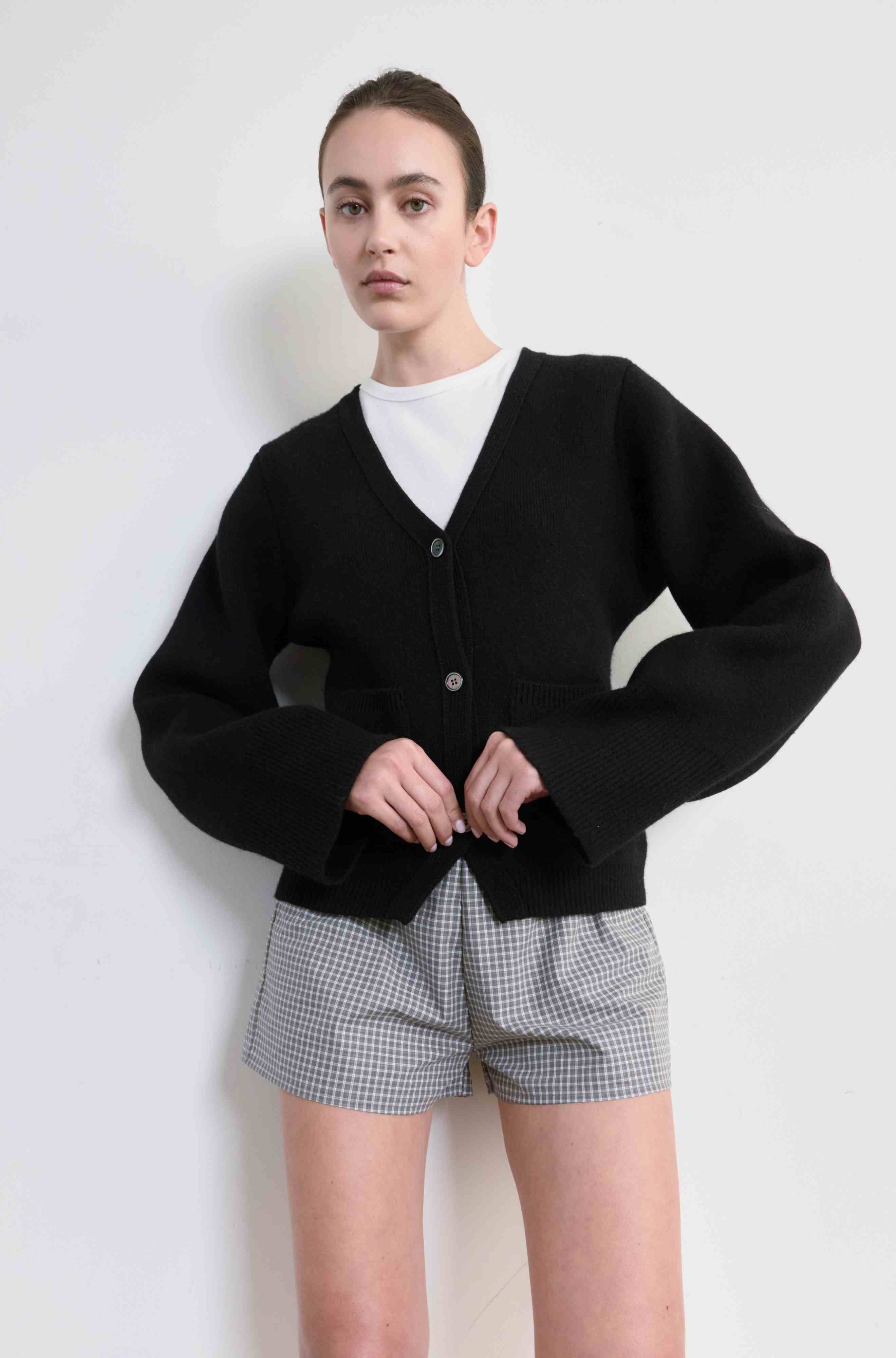Person wearing a black cardigan over a white shirt and plaid shorts on a light background
