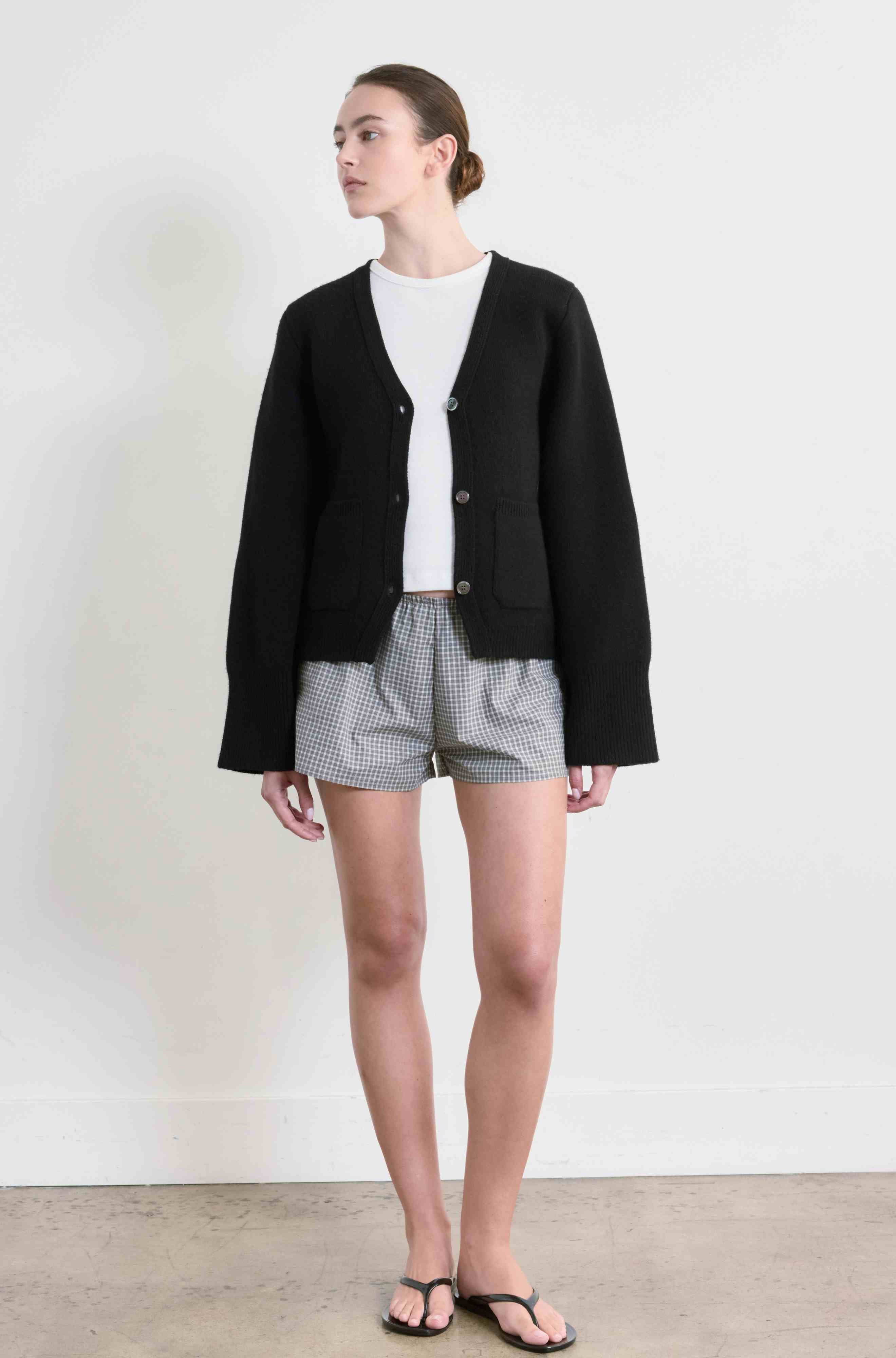 Woman wearing a black cardigan, white shirt, and checkered shorts on a plain background