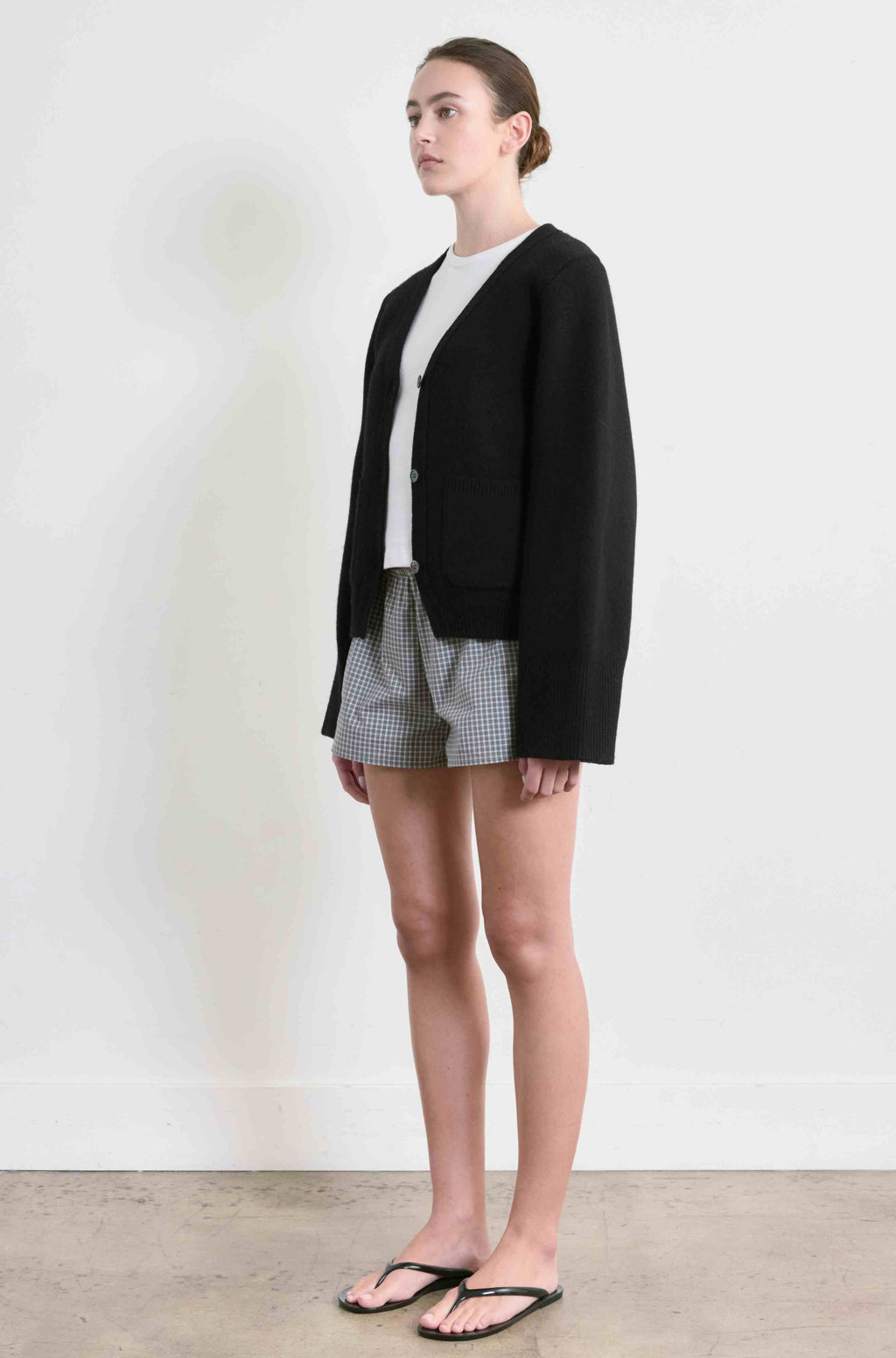 Woman wearing a black cardigan over a white shirt and checkered shorts on a plain background