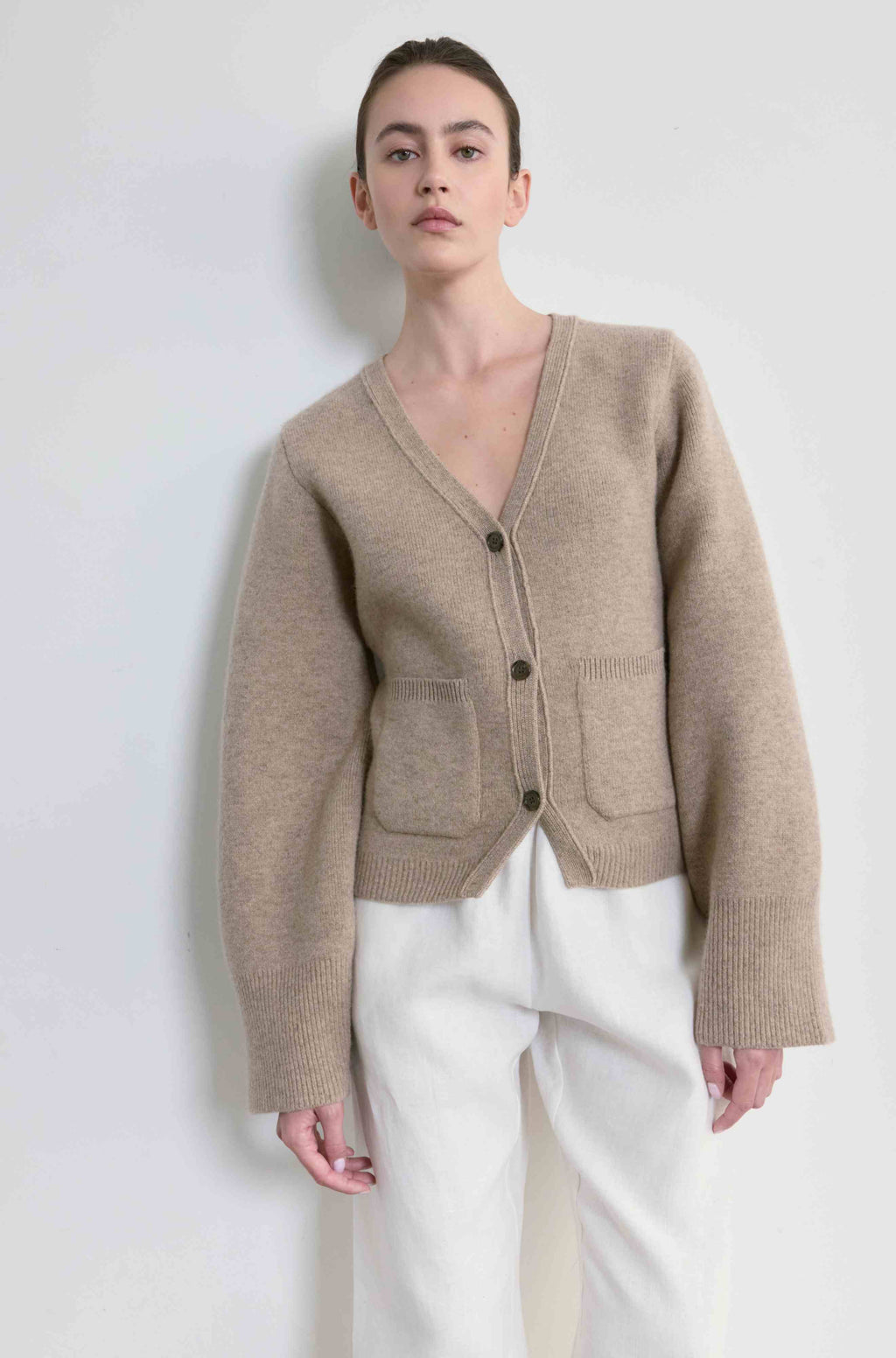 Person wearing a beige cardigan and white pants on a plain background