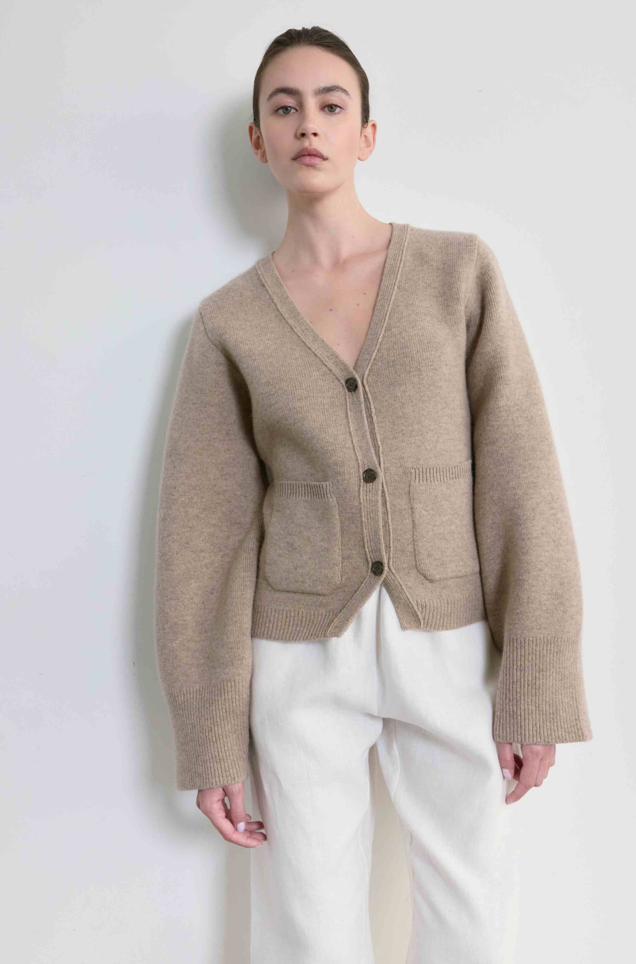 Person wearing a beige cardigan and white pants on a plain background