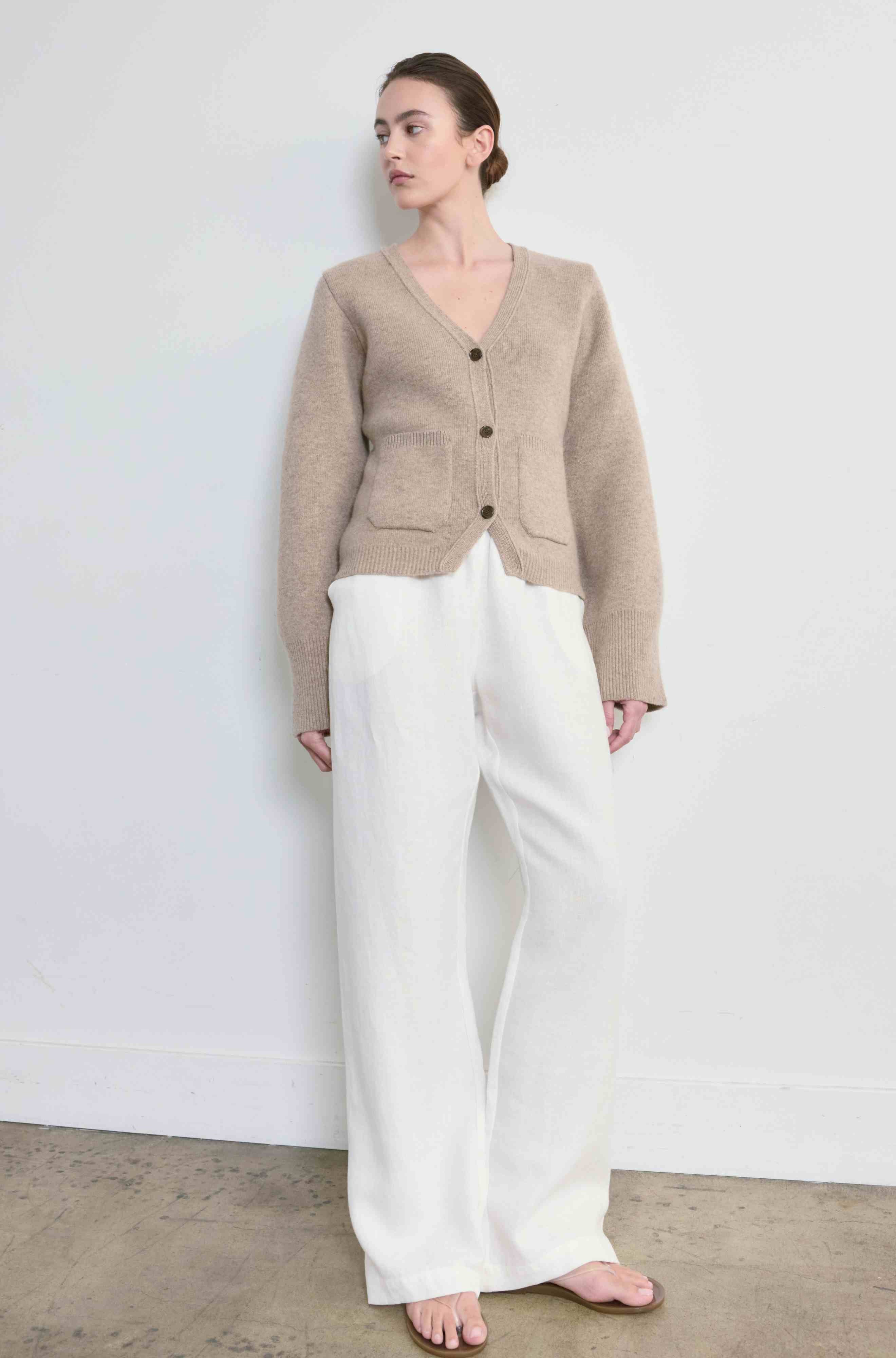 Woman wearing a beige cardigan and white pants against a plain background