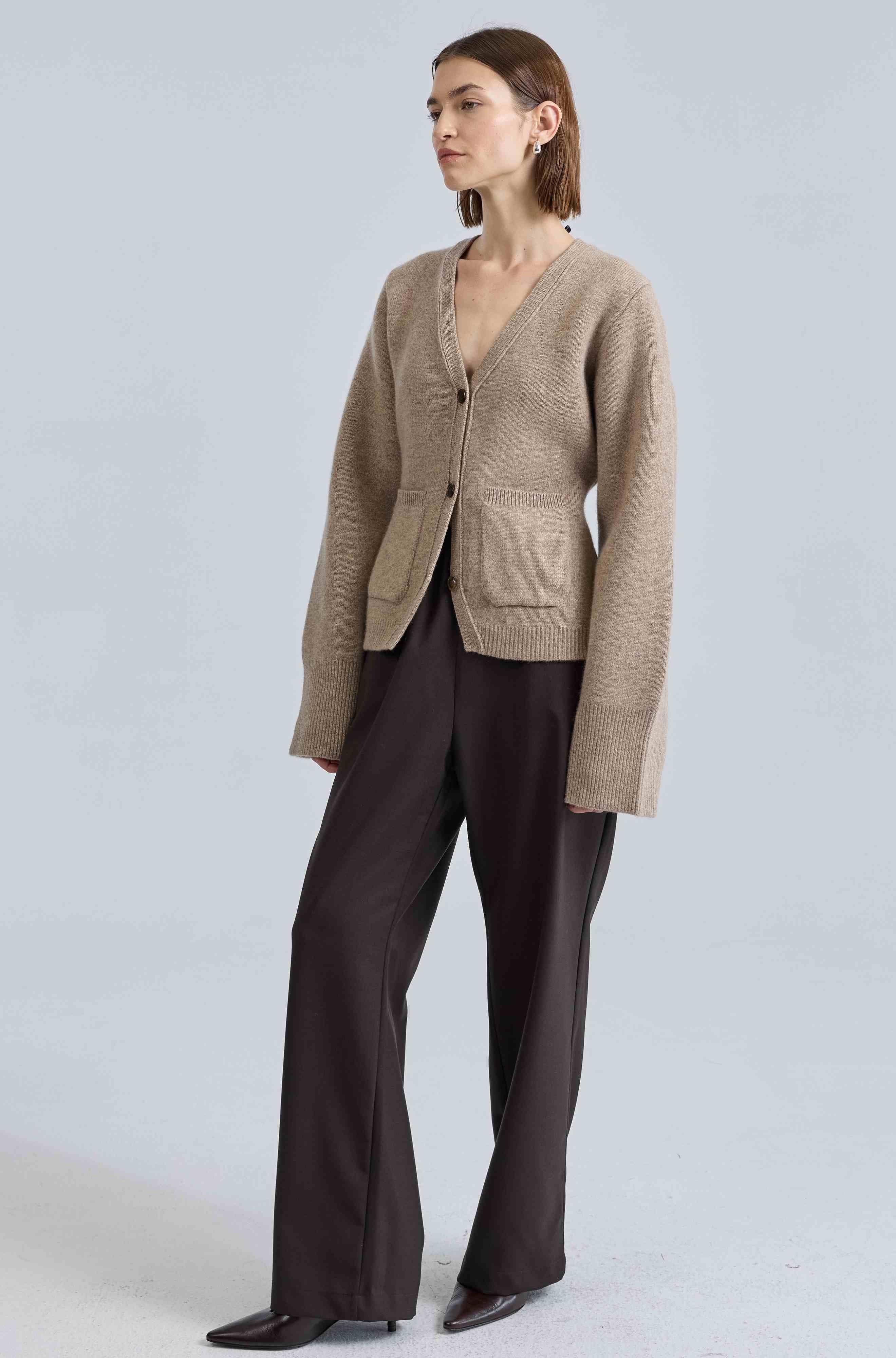 Woman wearing a beige cardigan and dark pants on a light gray background