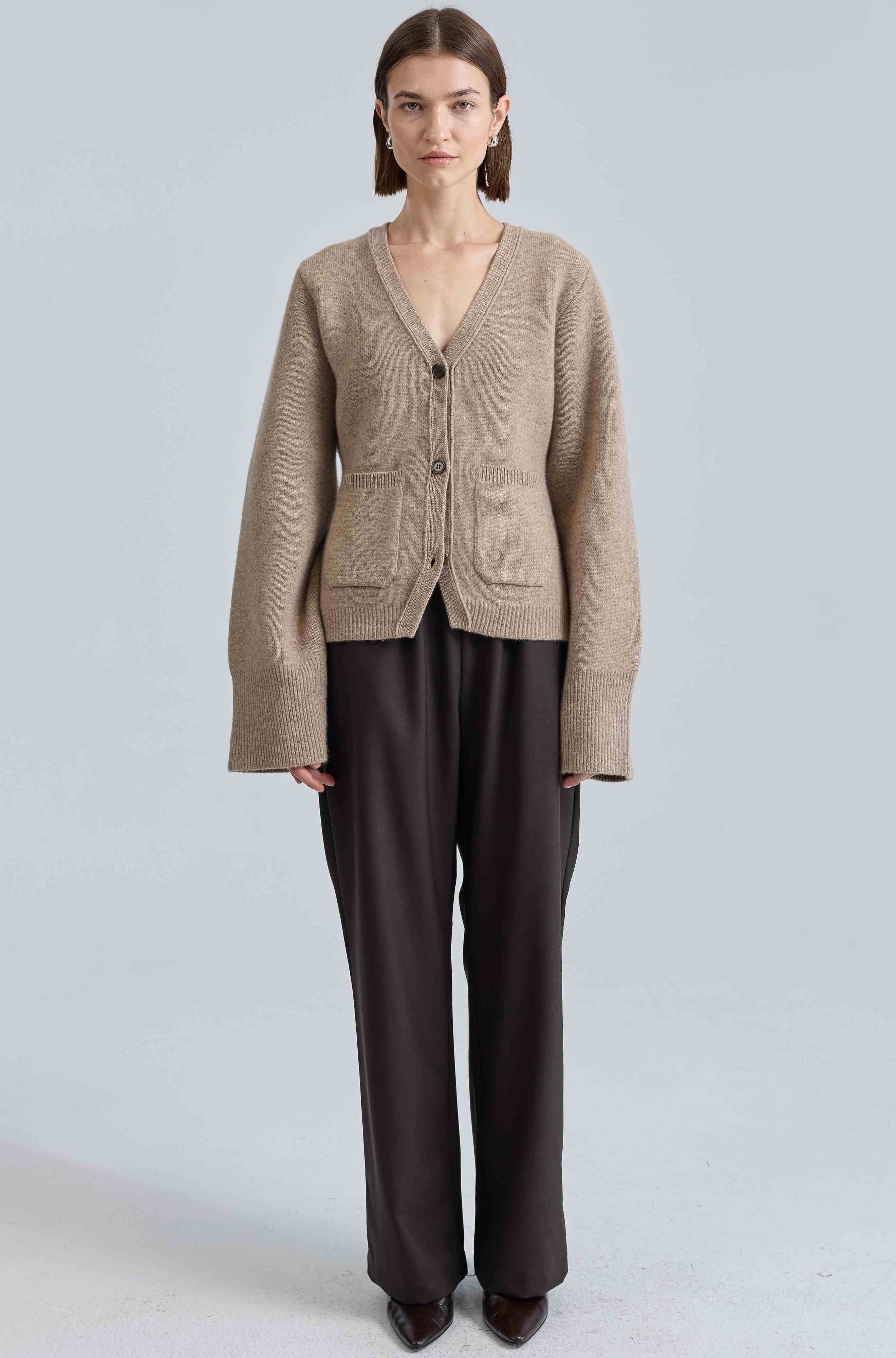 Woman wearing a beige cardigan and dark pants on a light gray background