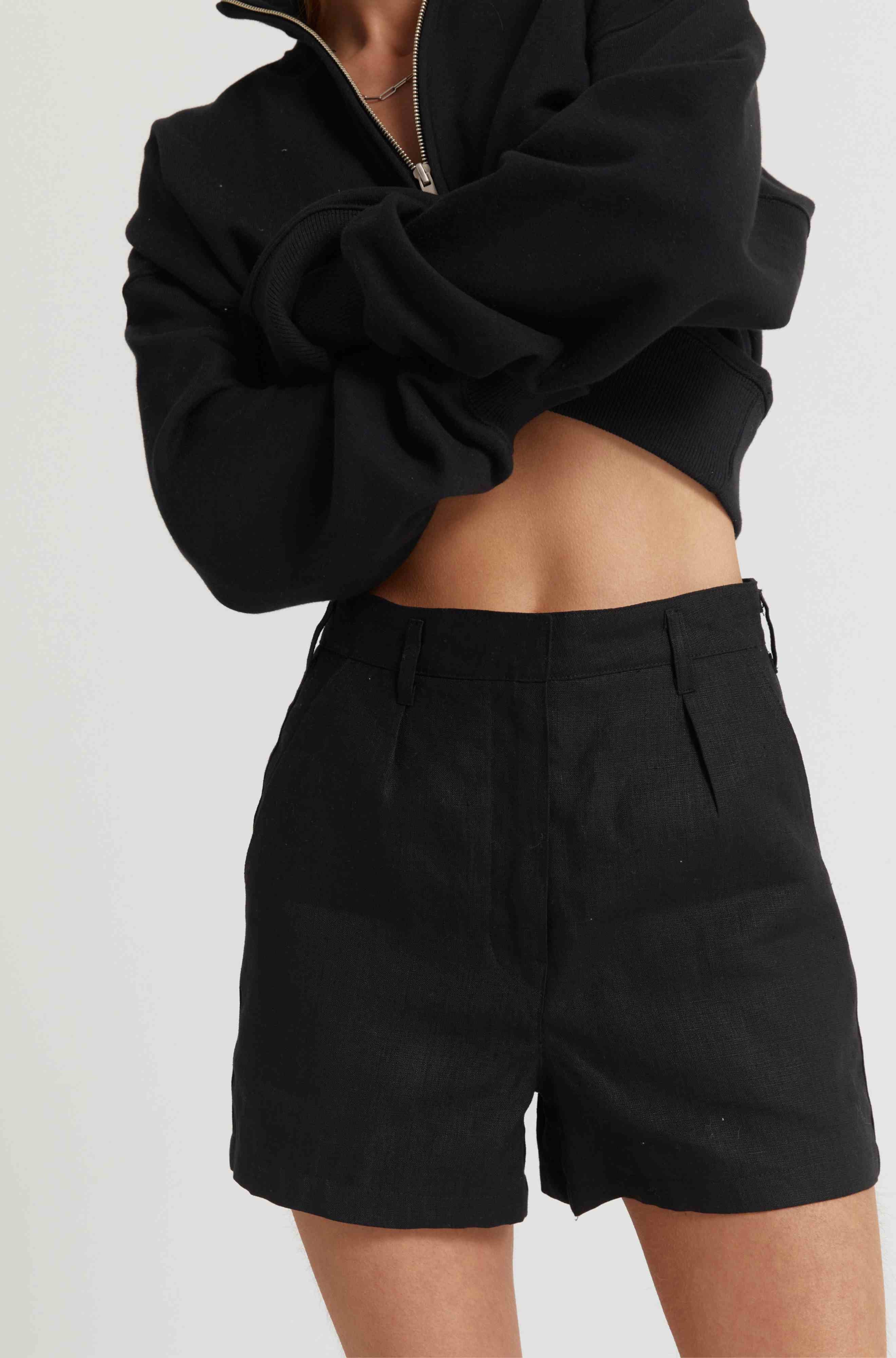 Woman wearing a black zip up sweatshirt and black linen shorts against a light grey background