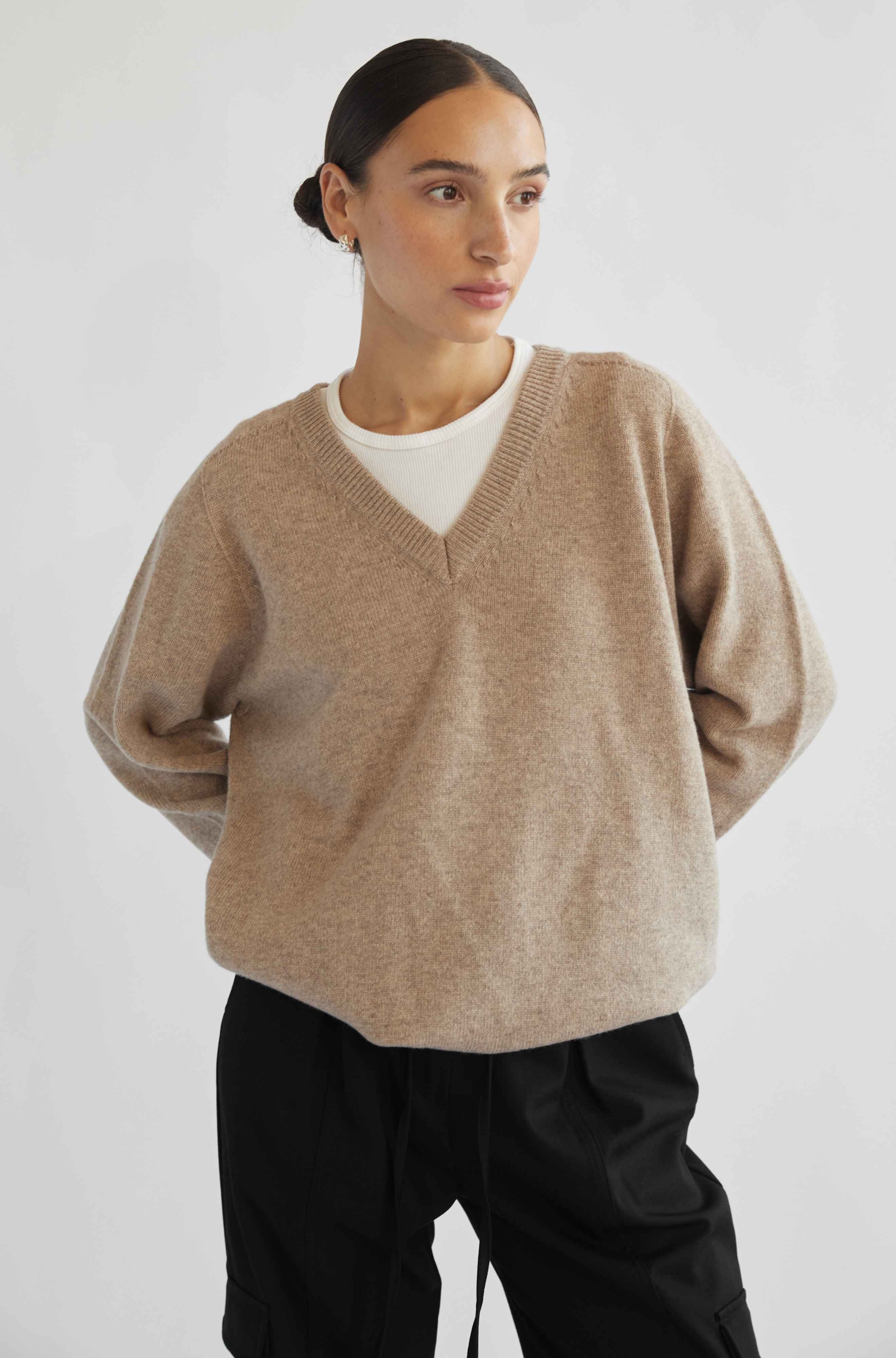 Woman wearing a beige sweater over a white shirt with black pants on a light gray background
