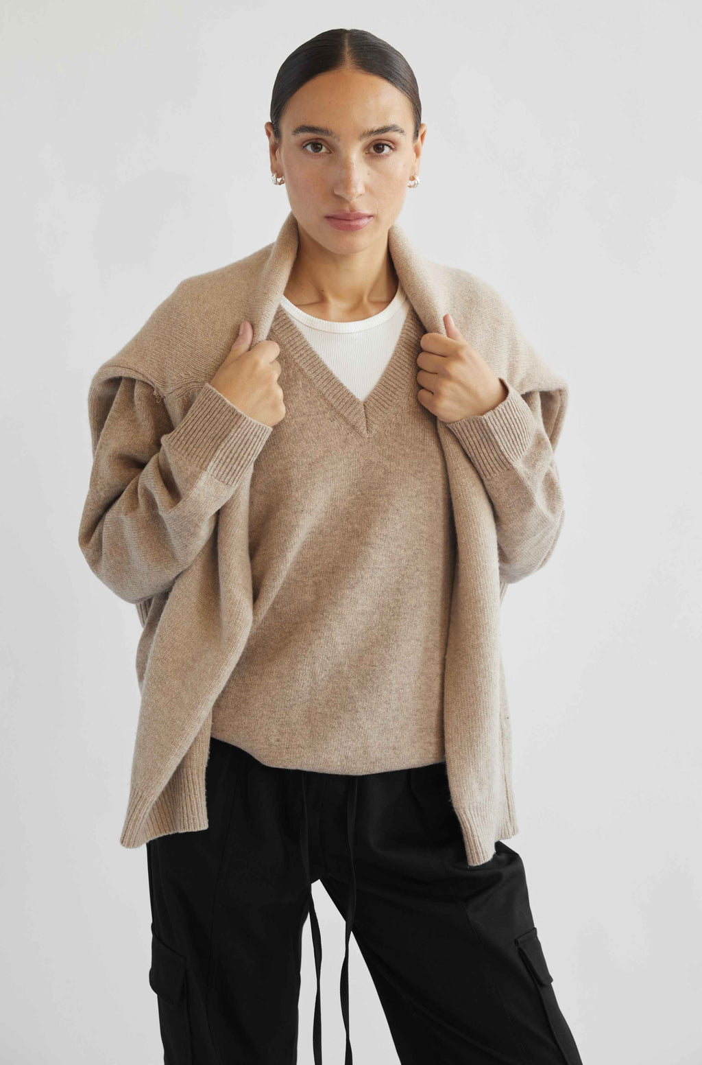 Woman wearing a beige cardigan over a white shirt with black pants on a white background