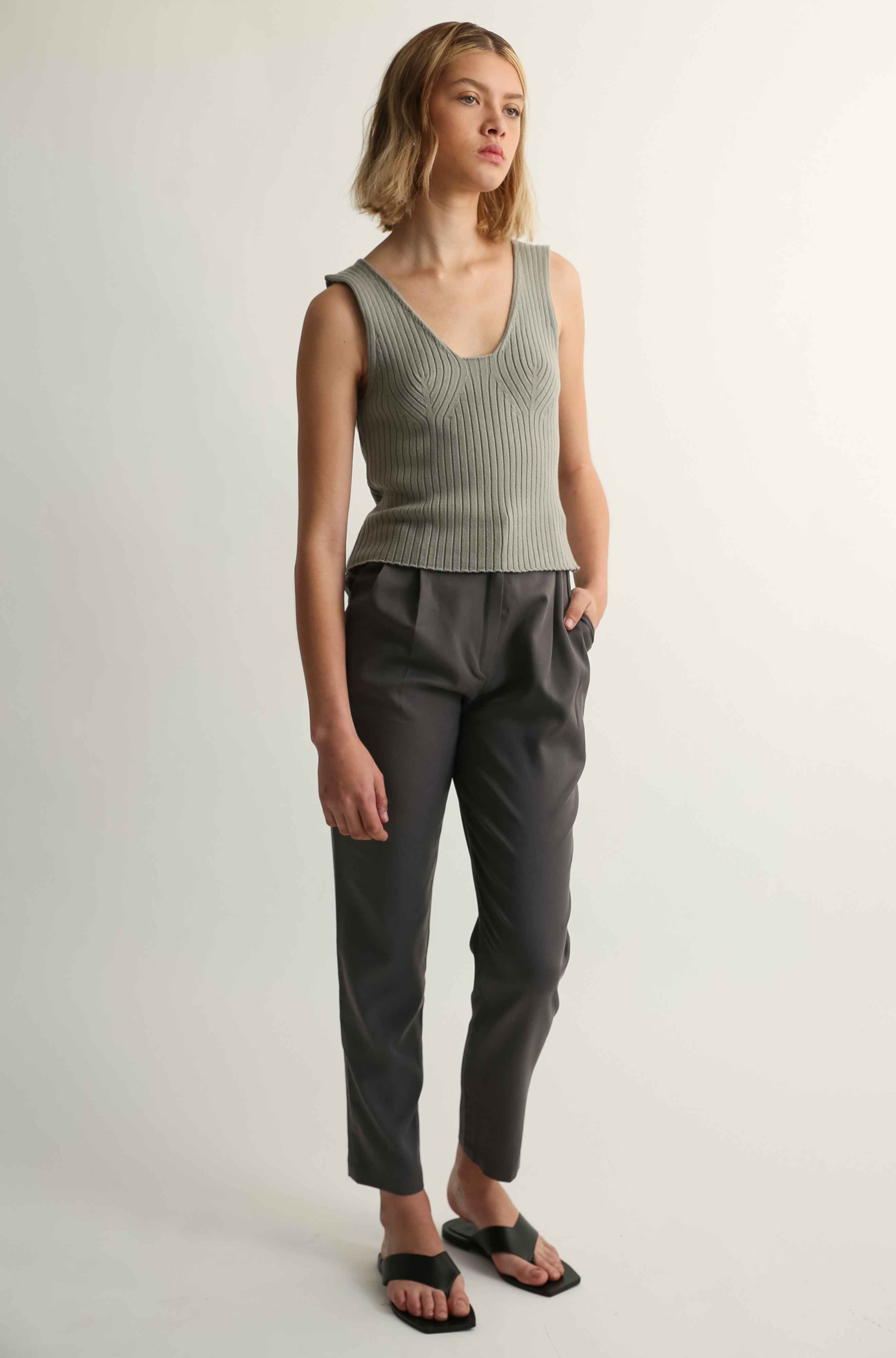Woman wearing a green ribbed tank top and dark pants on a plain background