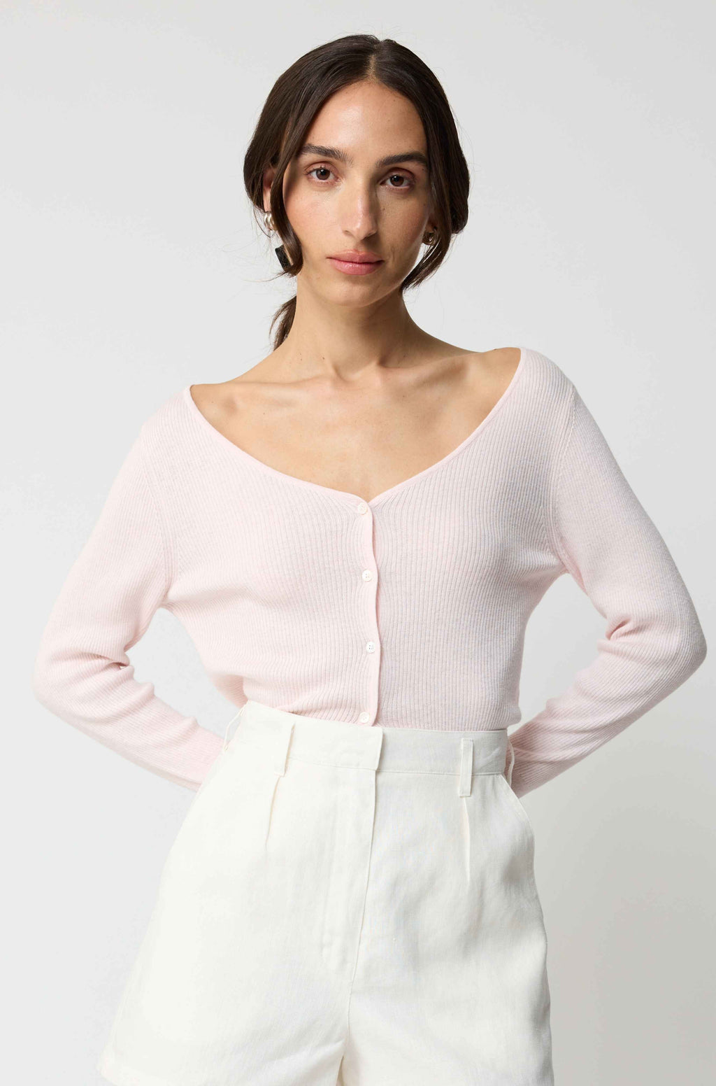 Woman wearing a light pink long-sleeve top and white pants on a plain background