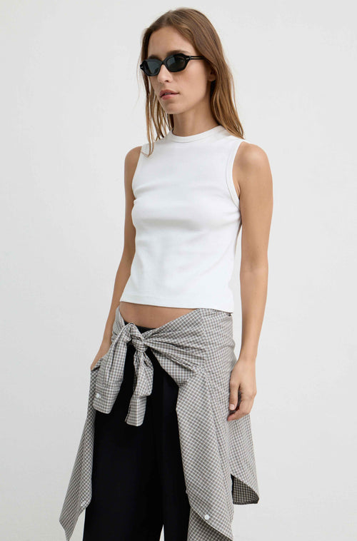 A woman modeling a white ribbed sleeveless mock-neck crop top, styled with a black-and-white gingham shirt tied around her waist and black pants, against a plain white studio background.