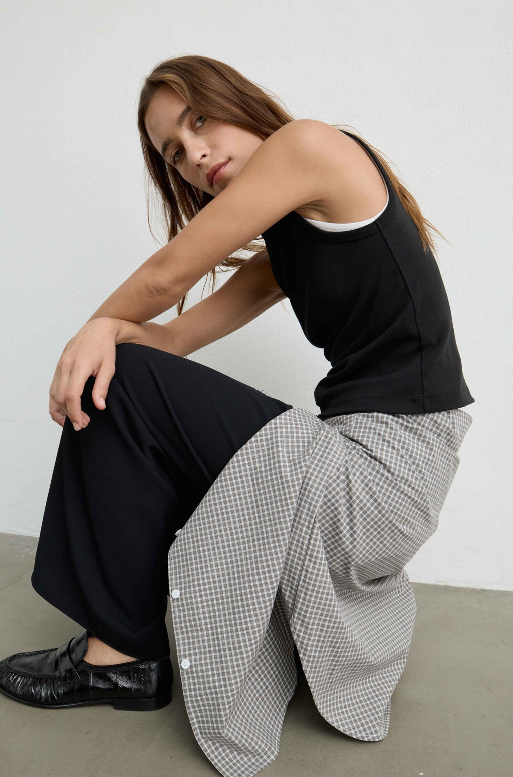 A woman crouches in a studio wearing a black tank top, wide-leg black trousers, and a gray-and-white gingham shirt draped over one leg, paired with black loafers.