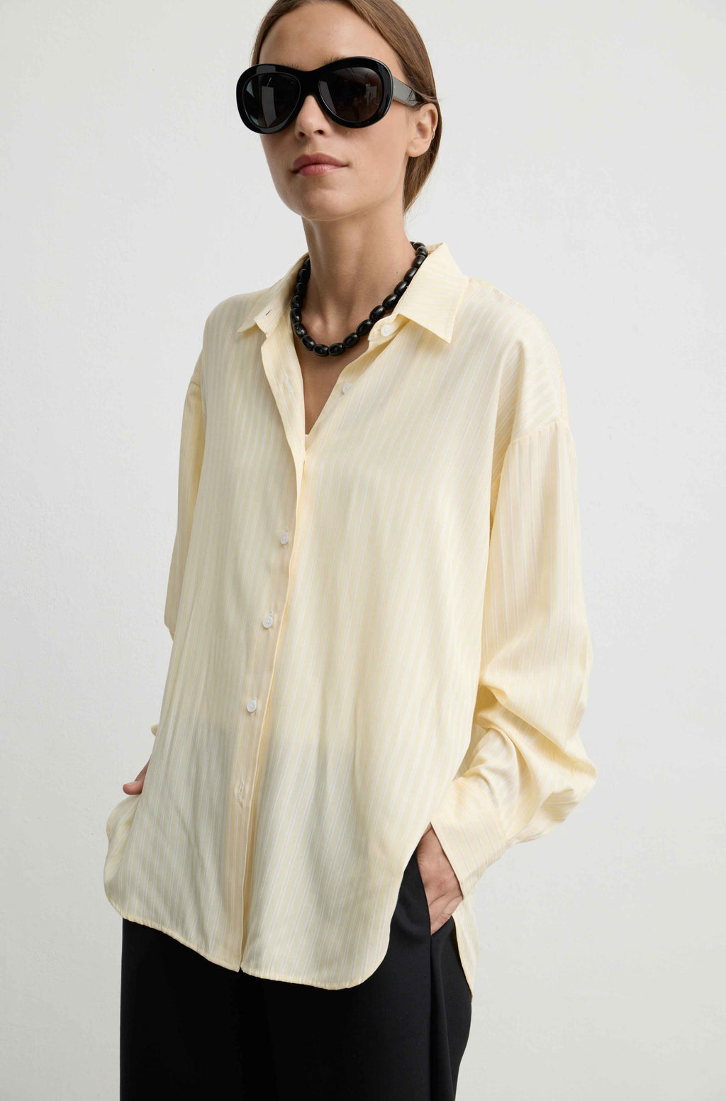 A woman wearing a pale yellow-colored, long-sleeved button-up shirt with subtle vertical stripes, styled with black pants, a chunky black beaded necklace, and oversized black sunglasses against a plain light gray background.