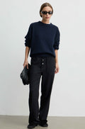 A model wearing a dark navy crewneck sweater, black wide-leg trousers, black sunglasses, and holding a black leather clutch bag, standing against a plain light gray studio background.